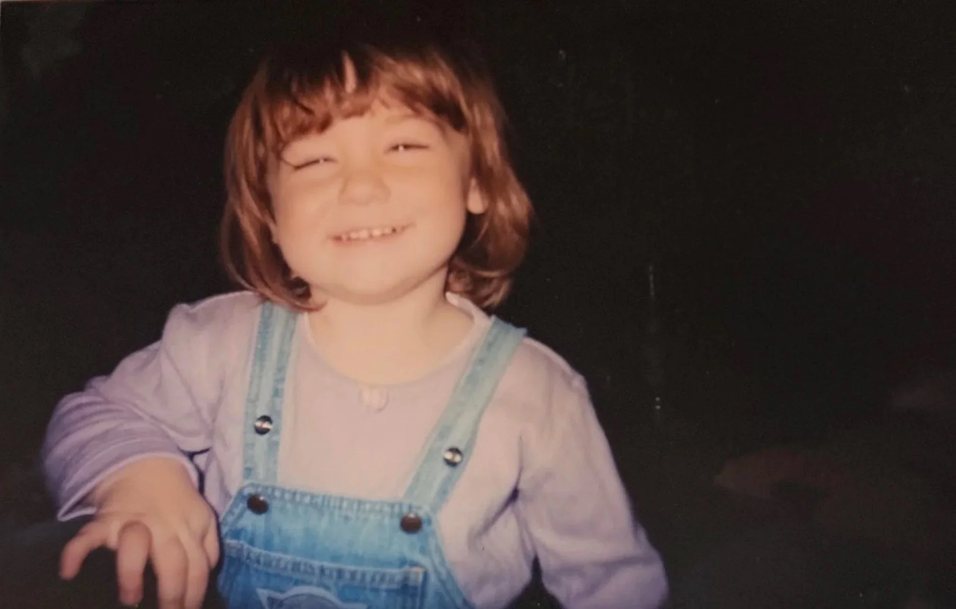 A young girl with shoulder-length brown hair smiling, wearing a light-colored shirt and denim overalls, in front of a dark background.