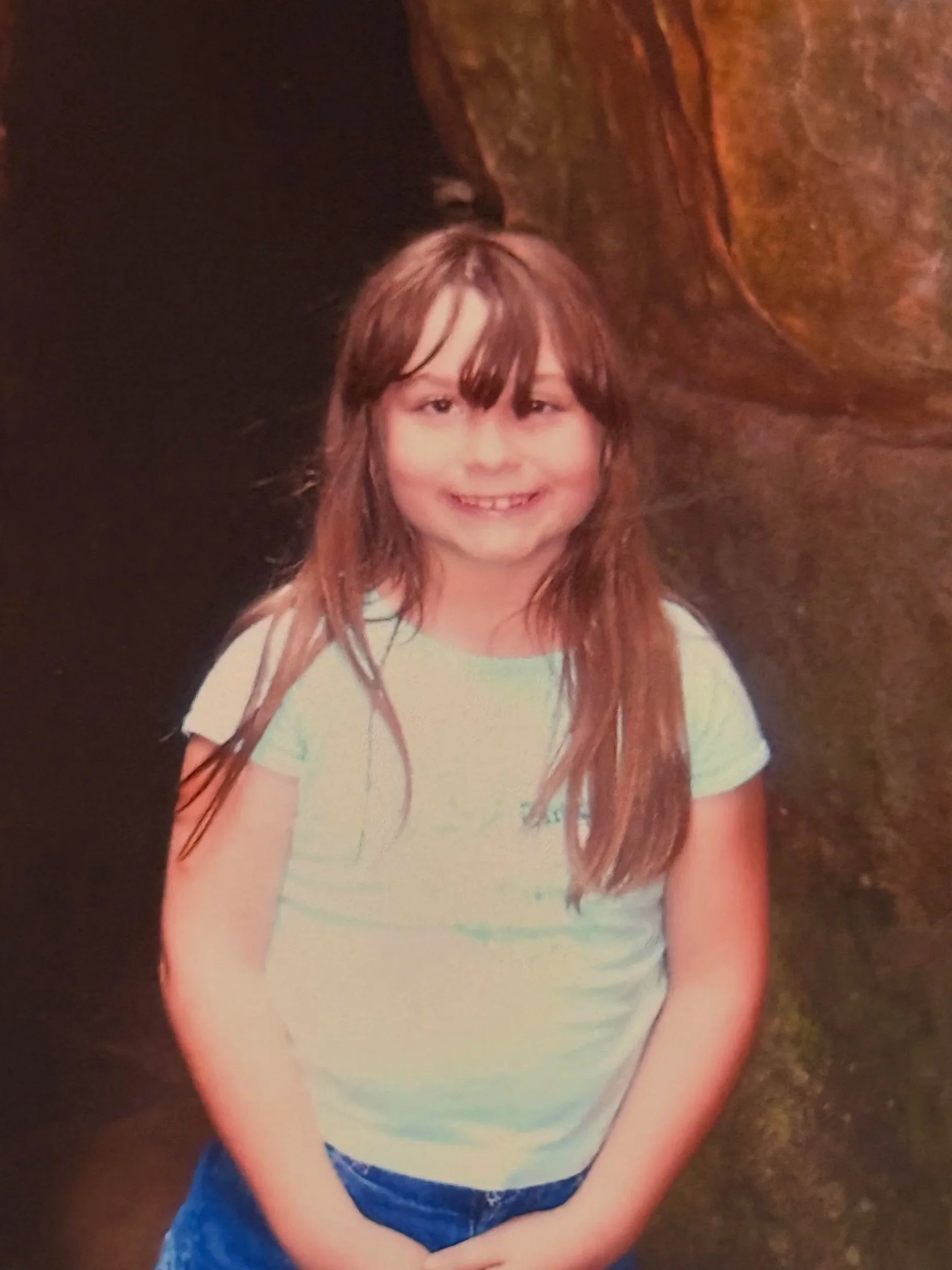 A young girl with long auburn hair and a fringe smiling with missing front teeth, wearing a light-colored t-shirt and blue jeans, standing outdoors on a dirt or gravel surface.