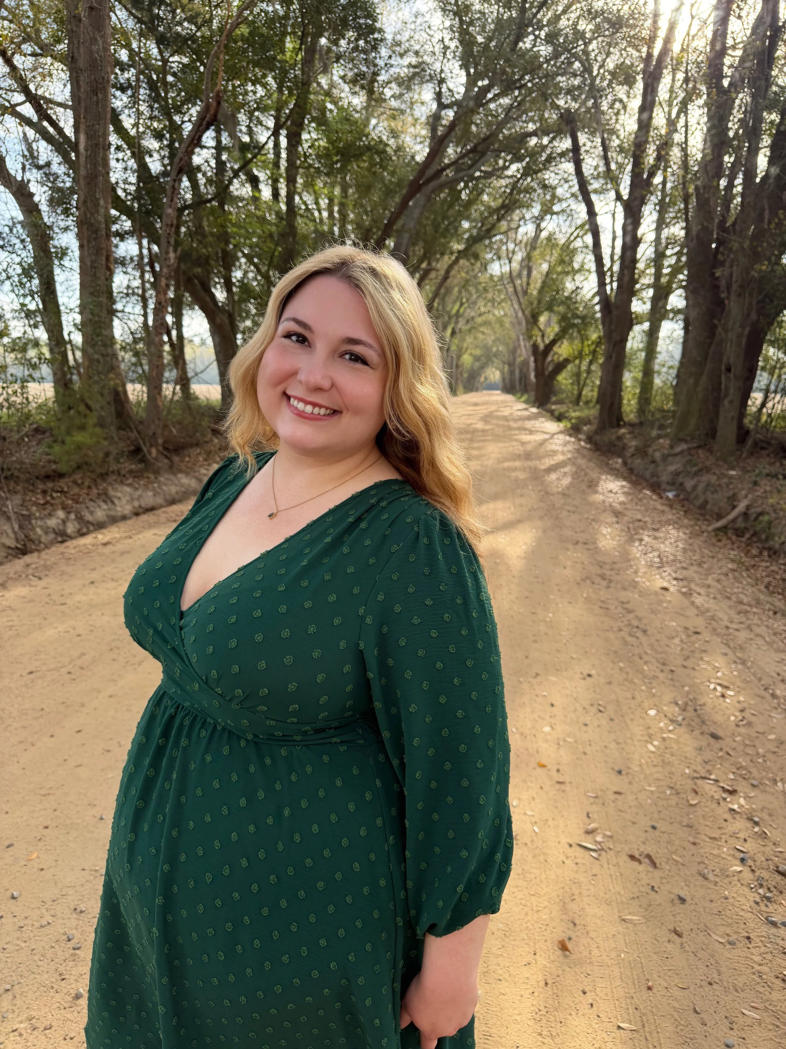 A smiling woman with blonde hair in a green dress standing on a dirt road lined with trees.