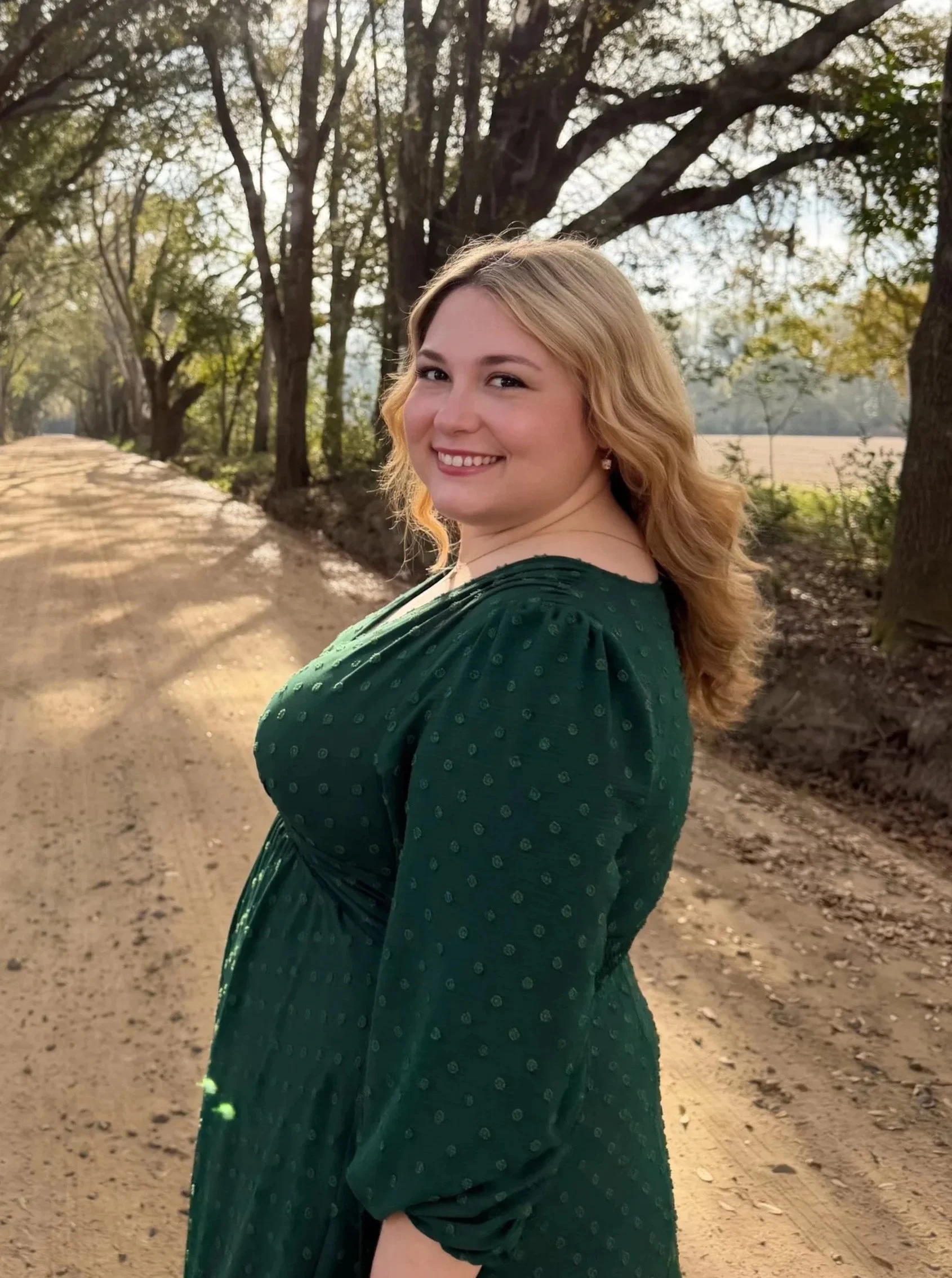 A woman with blonde hair smiling while standing on a dirt path lined with trees during a sunny day.
