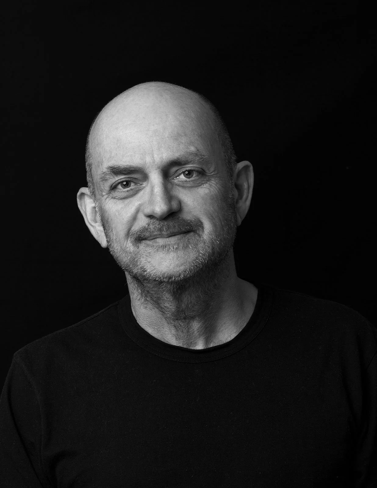 Black and white portrait of a middle-aged man with a beard, bald head, wearing a black shirt, smiling slightly, against a black background.