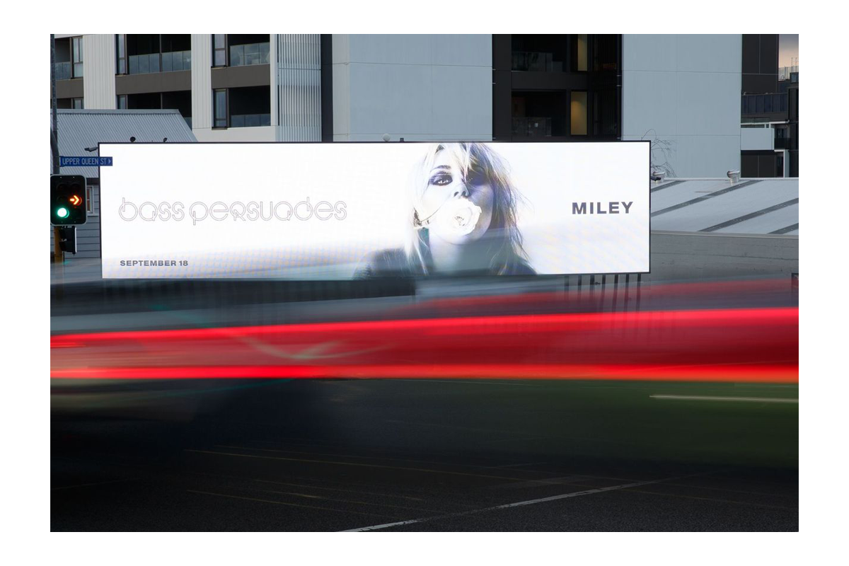 Billboard advertisement for Miley Cyrus's album 'Bangerz' showing a woman blowing a bubblegum bubble, set against a city street with a green traffic light and streaks of red and white lights from passing cars.