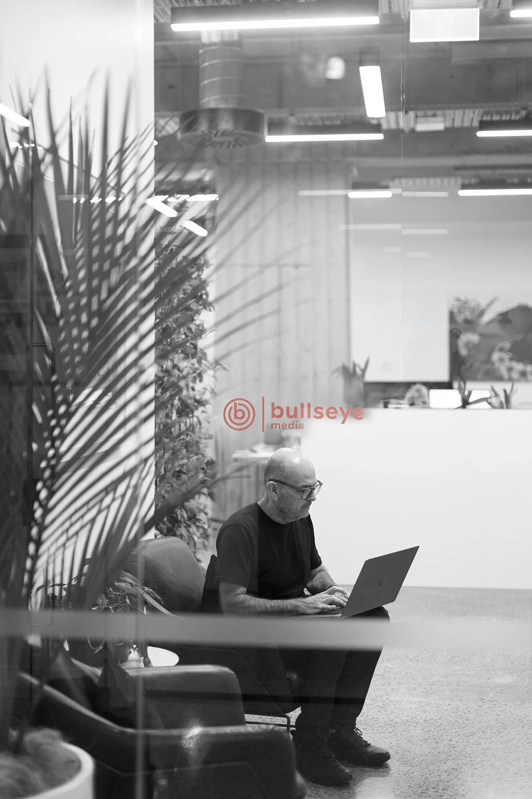A black and white photo of a man working on a laptop in a modern office with plants and glass walls, seen through a window with the logo 'bullseye media' on it.