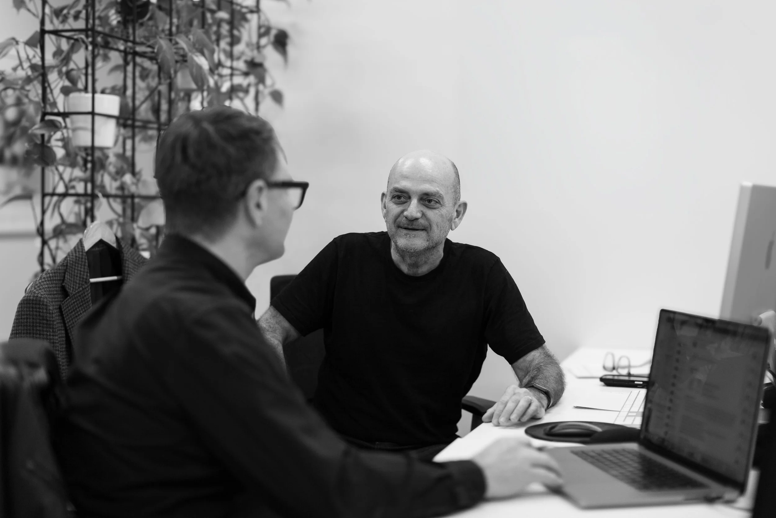 Two men having a conversation in an office, with a laptop on the desk and a plant in the background.
