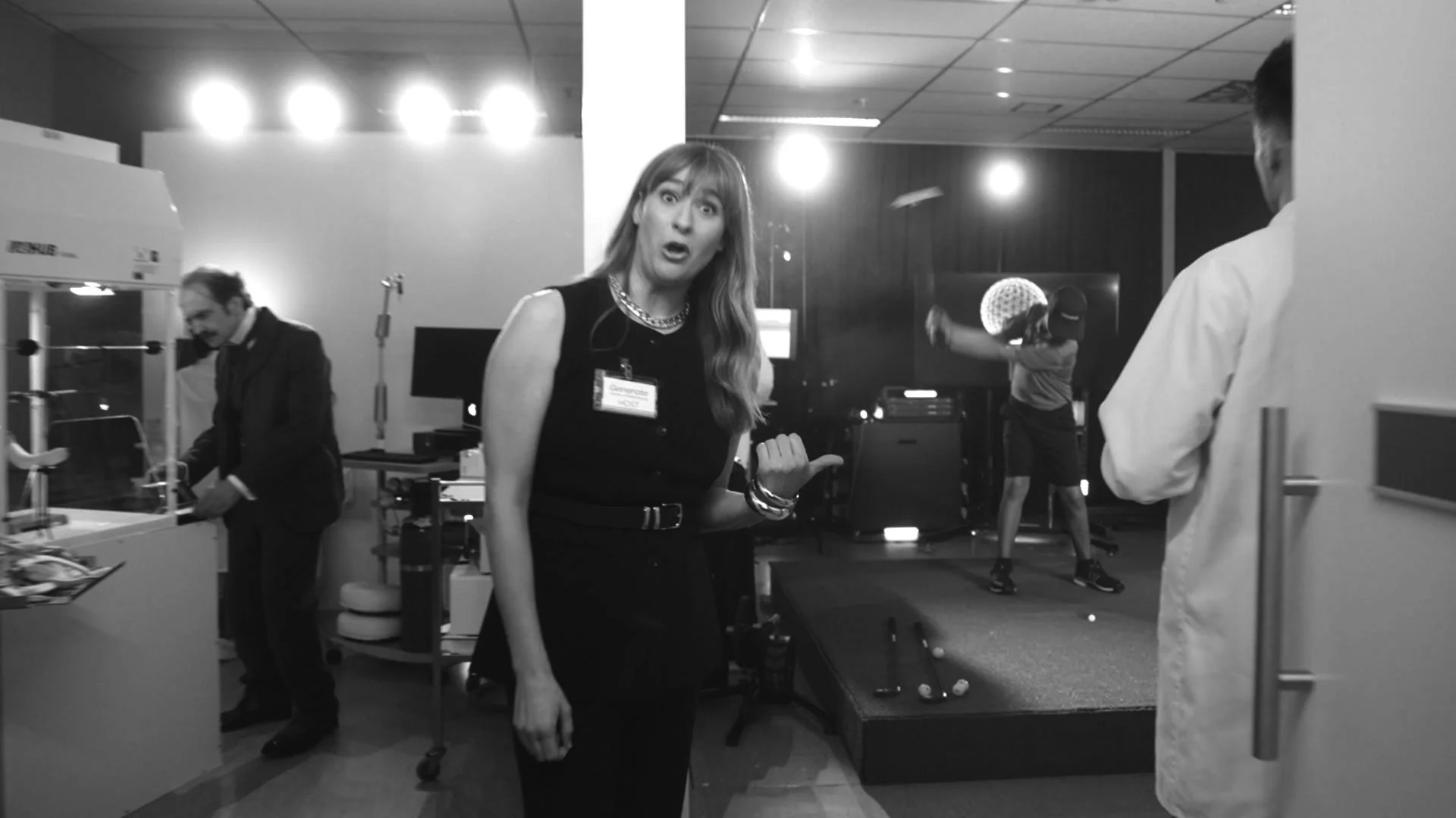 A woman with long hair and a dress, pointing her thumb towards herself, at a professional event or conference, with several people and equipment in the background.