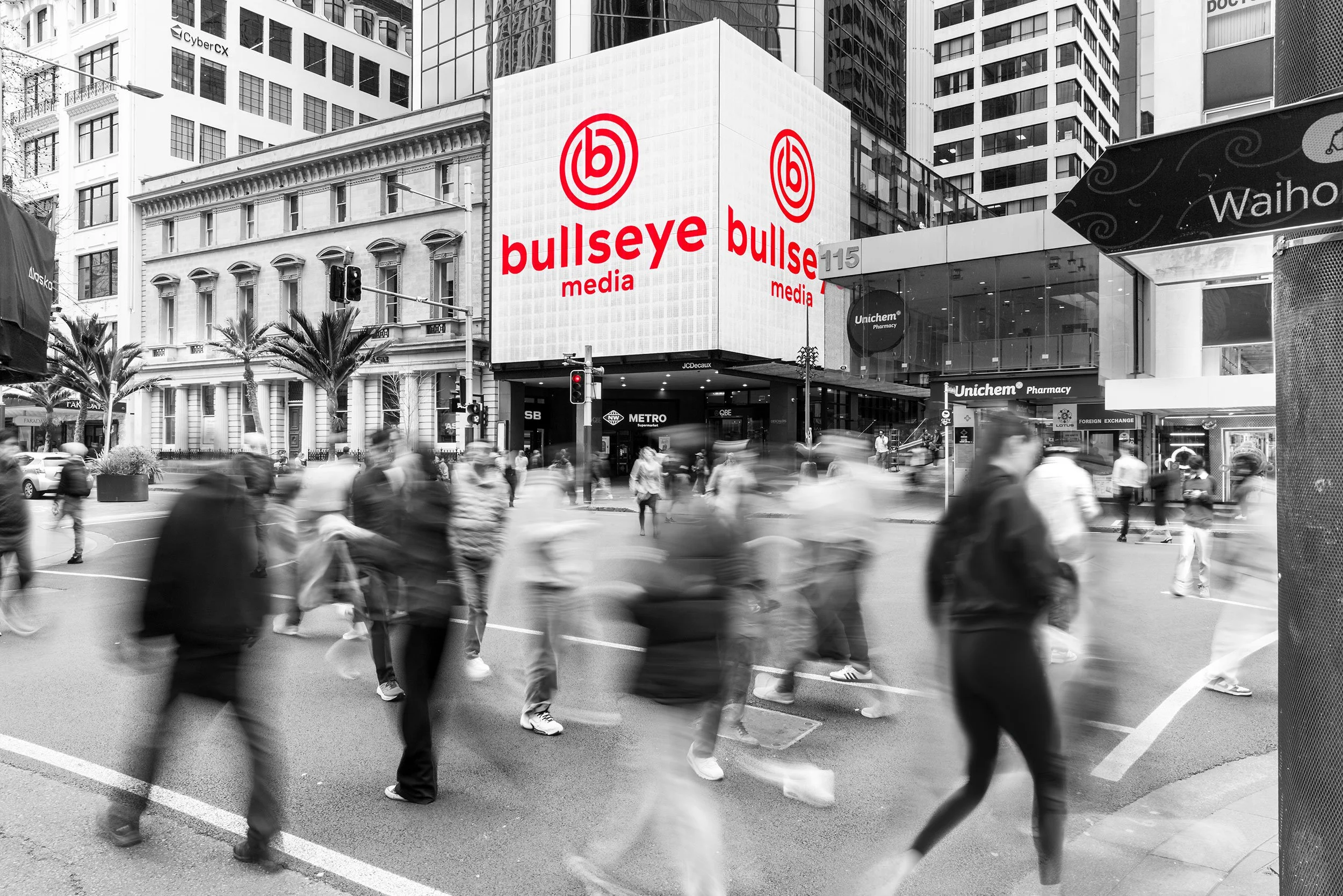 A busy city intersection with many pedestrians crossing the street in front of tall buildings. Prominent digital billboard displays the logo of Bullseye Media, with other storefronts visible, including a Unichem Pharmacy.