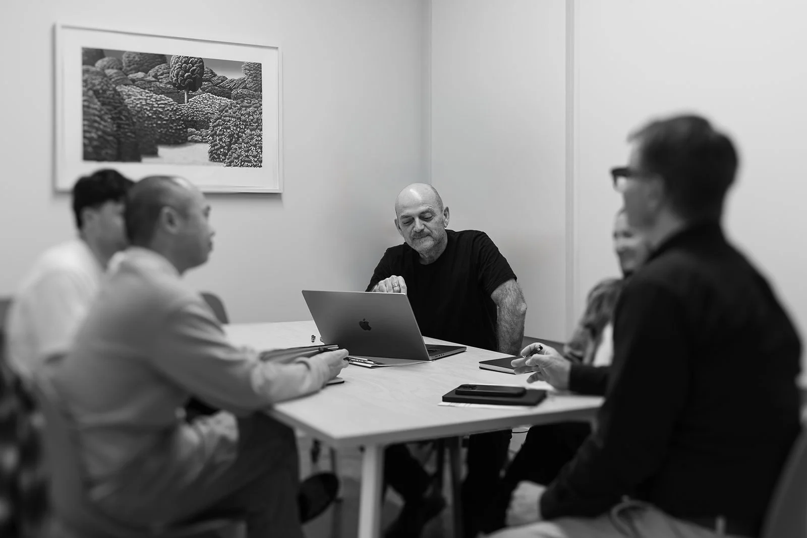A group of people in a meeting room, with four men visible, sitting around a table and talking. One man, with a bald head and gray beard, is sitting at the head of the table with a laptop, while others have notebooks and phones.
