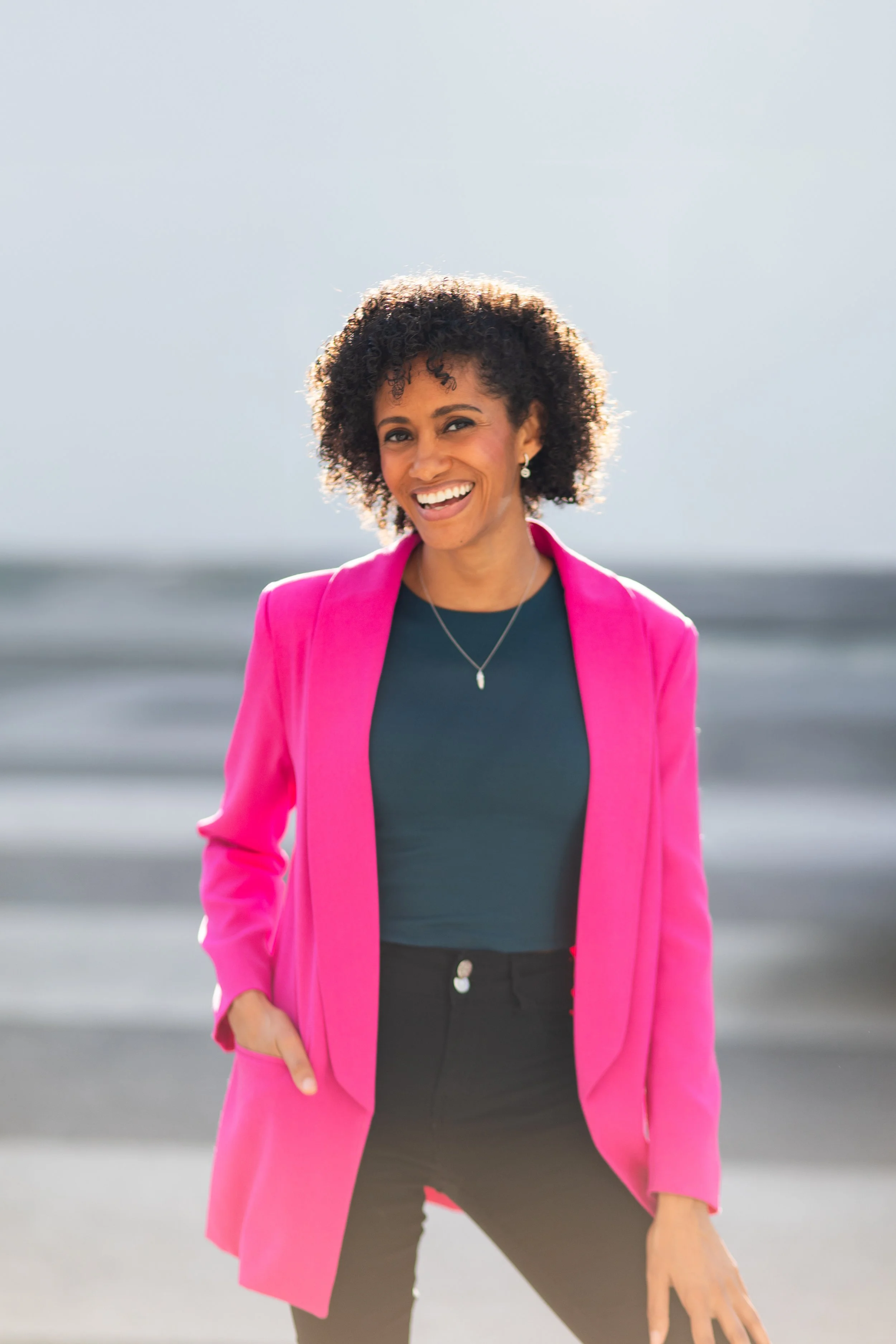 Photo of Crystal, a Black woman with short curly dark brown hair, wearing a dark teal top and barbie pink blazer, smiling at the camera