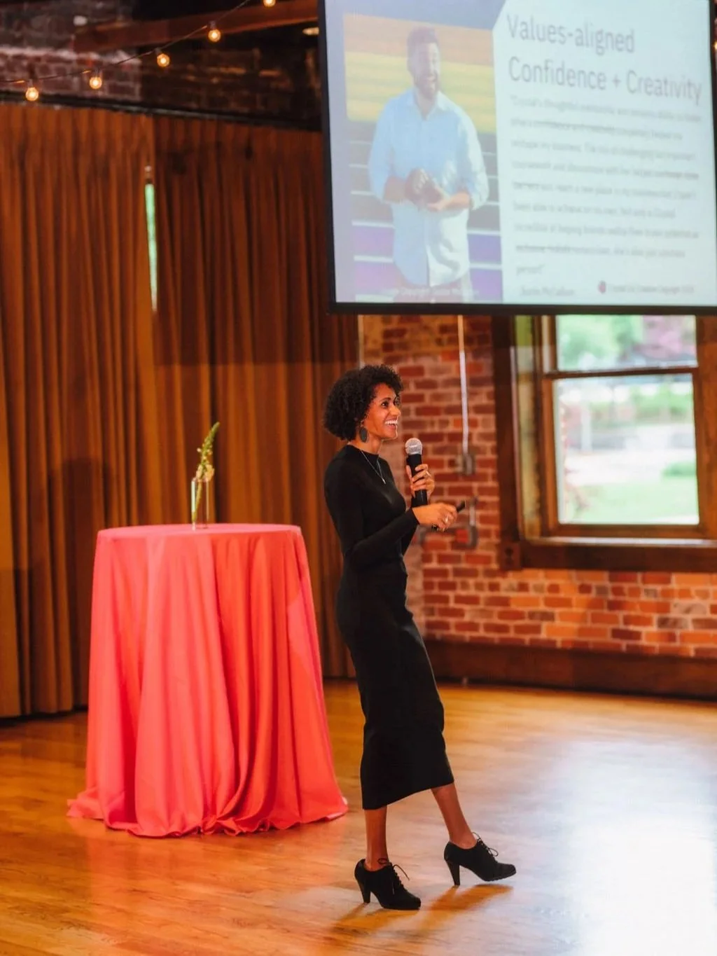 Photo of Crystal, a Black woman with shoulder length dark brown curly hair, wearing a black dress and black heels, holding a microphone, smiling while she speaks