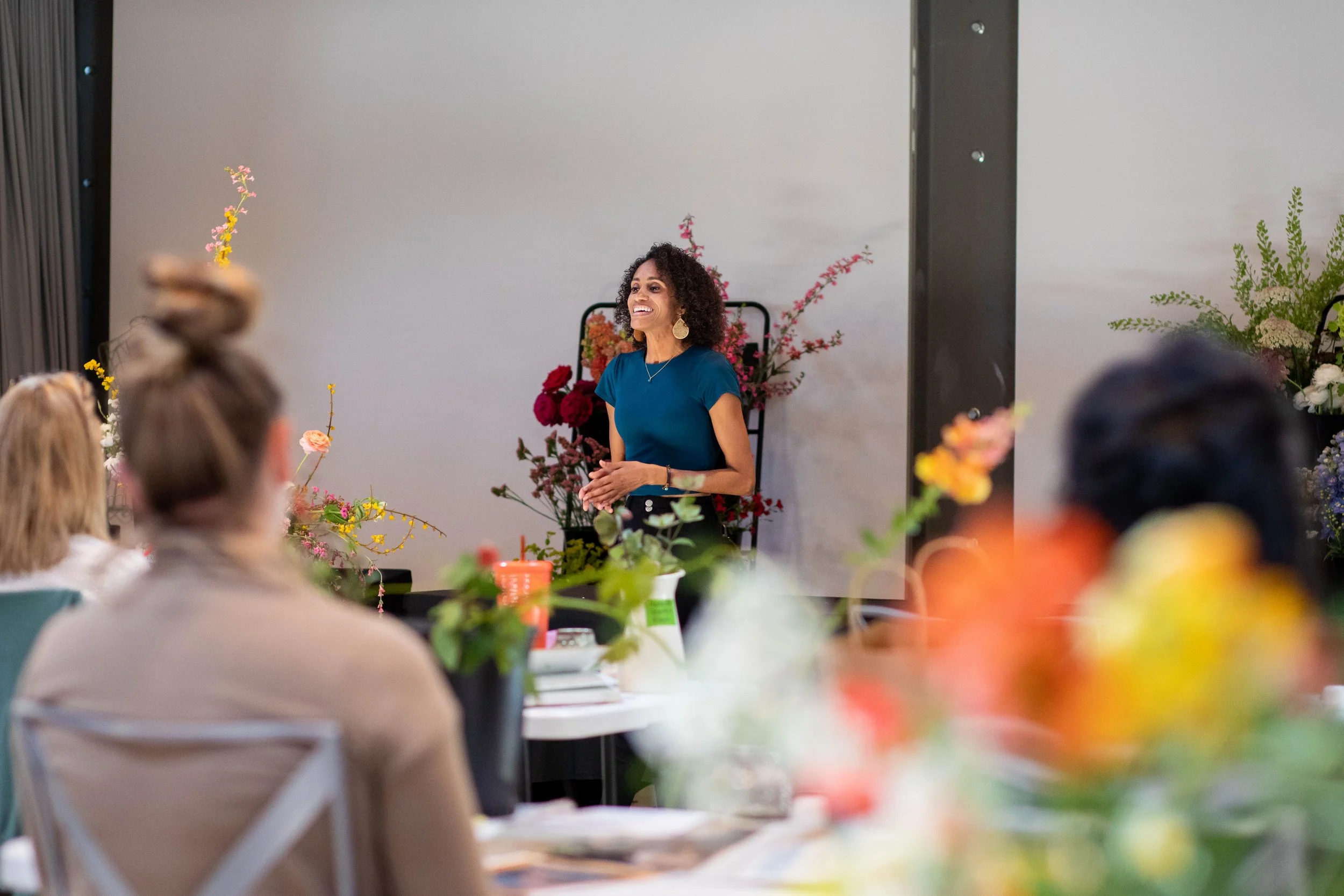 Photo of Crystal, a Black woman with shoulder length curly hair, standing in the front of a room, speaking, smiling at the attendees