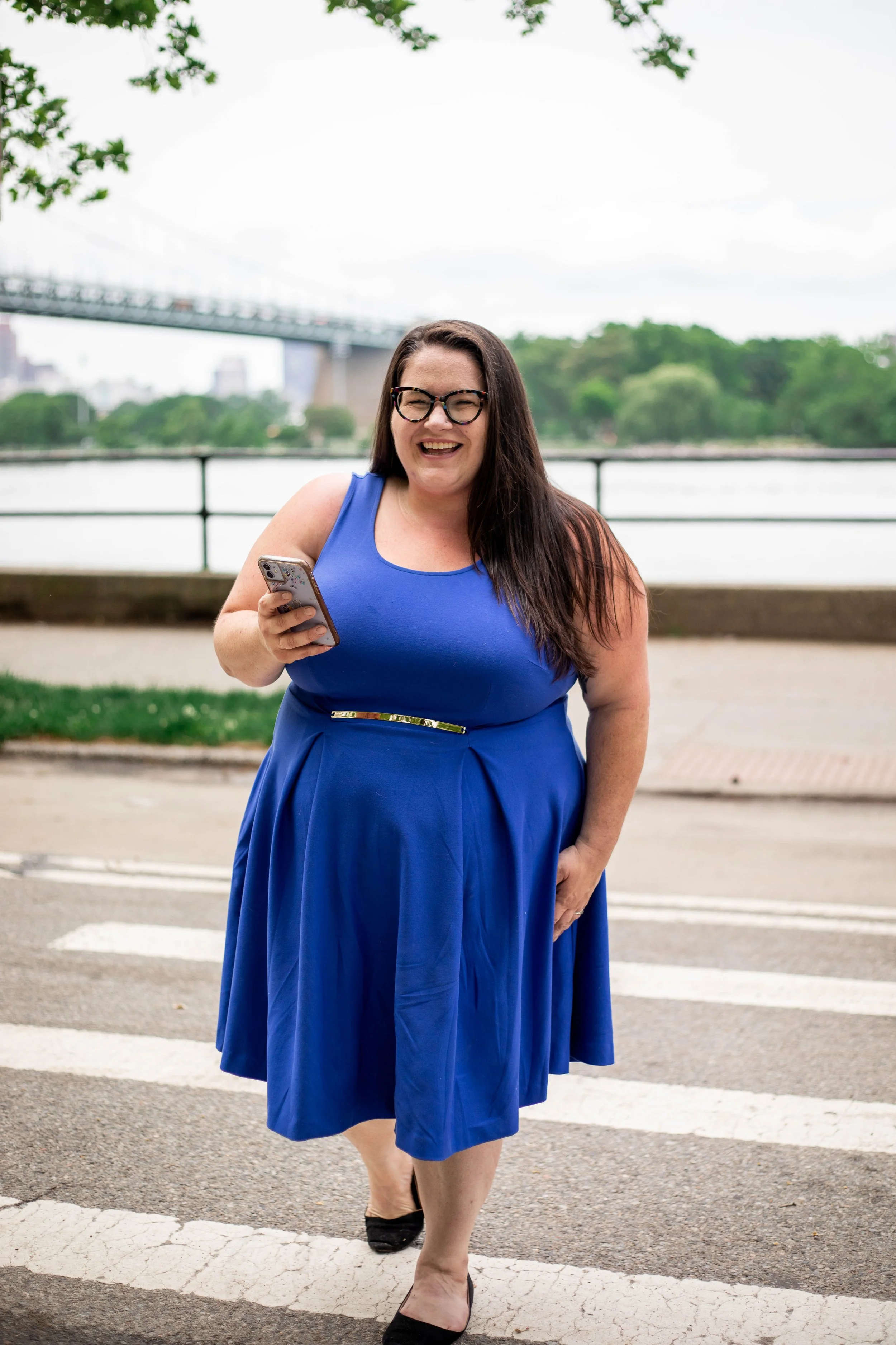 Photo of a plus size, white woman with long brown hair, wearing a blue dress, holding her phone, walking across a cross walk, smiling at the camera