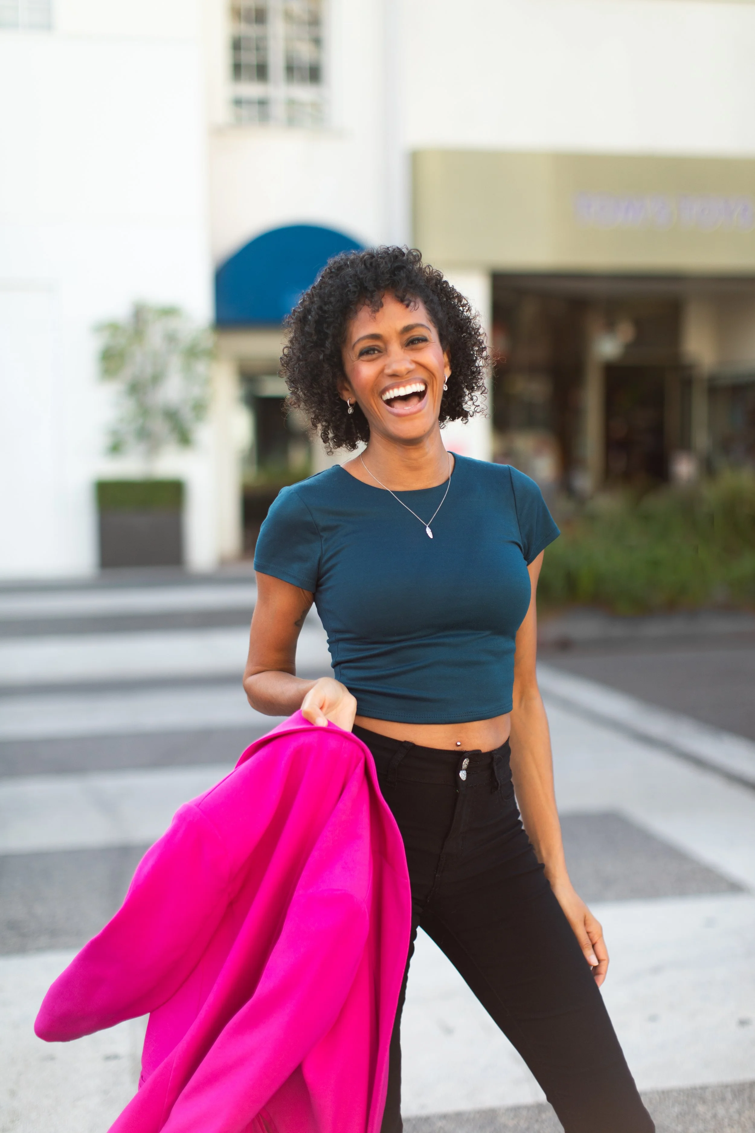 Photo of Crystal, a Black woman with short curly dark brown hair, wearing a dark teal top and holding a barbie pink blazer, smiling and laughing at the camera