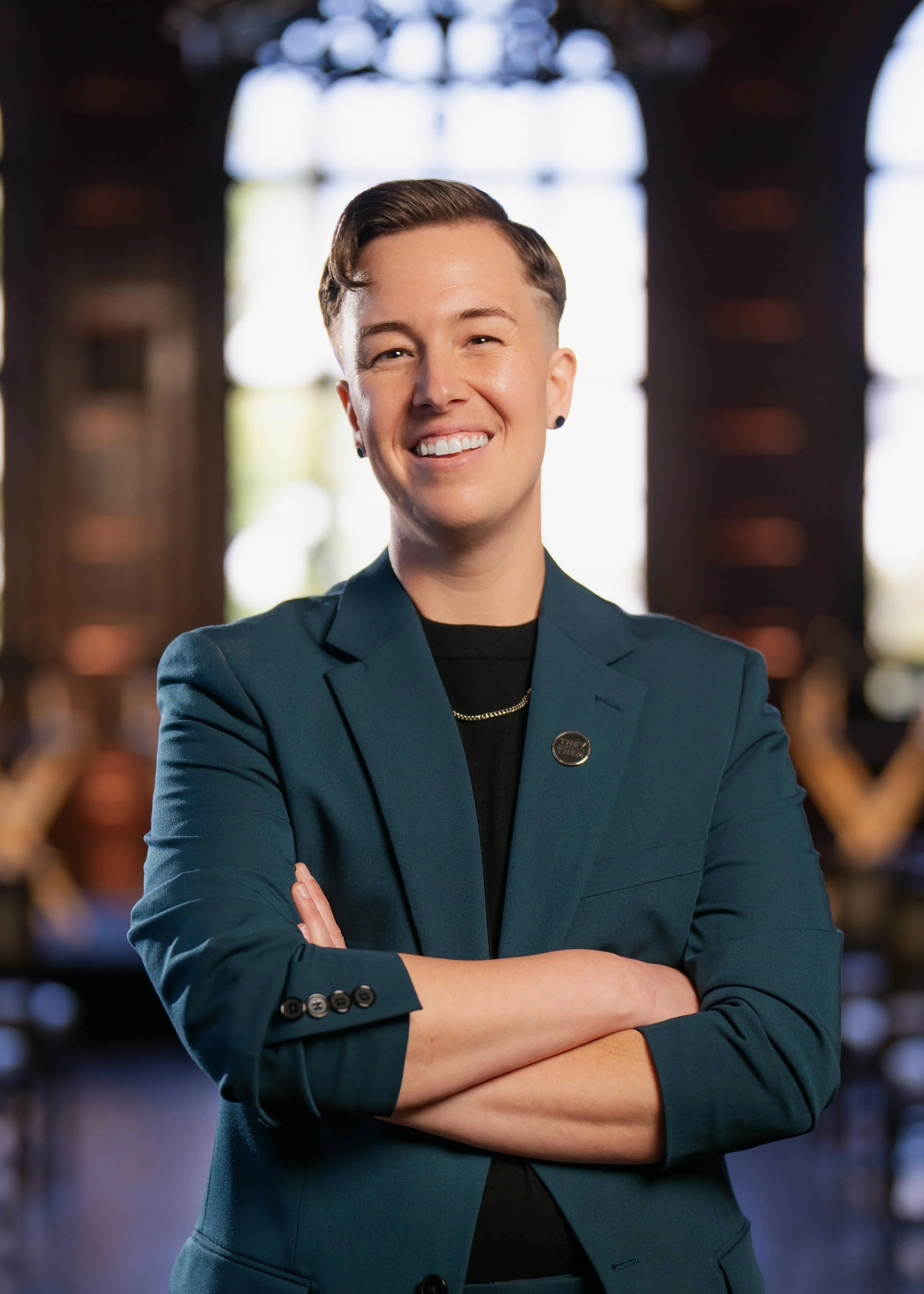 Headshot of a white, nonbinary person wearing a teal blazer, smiling at the camera