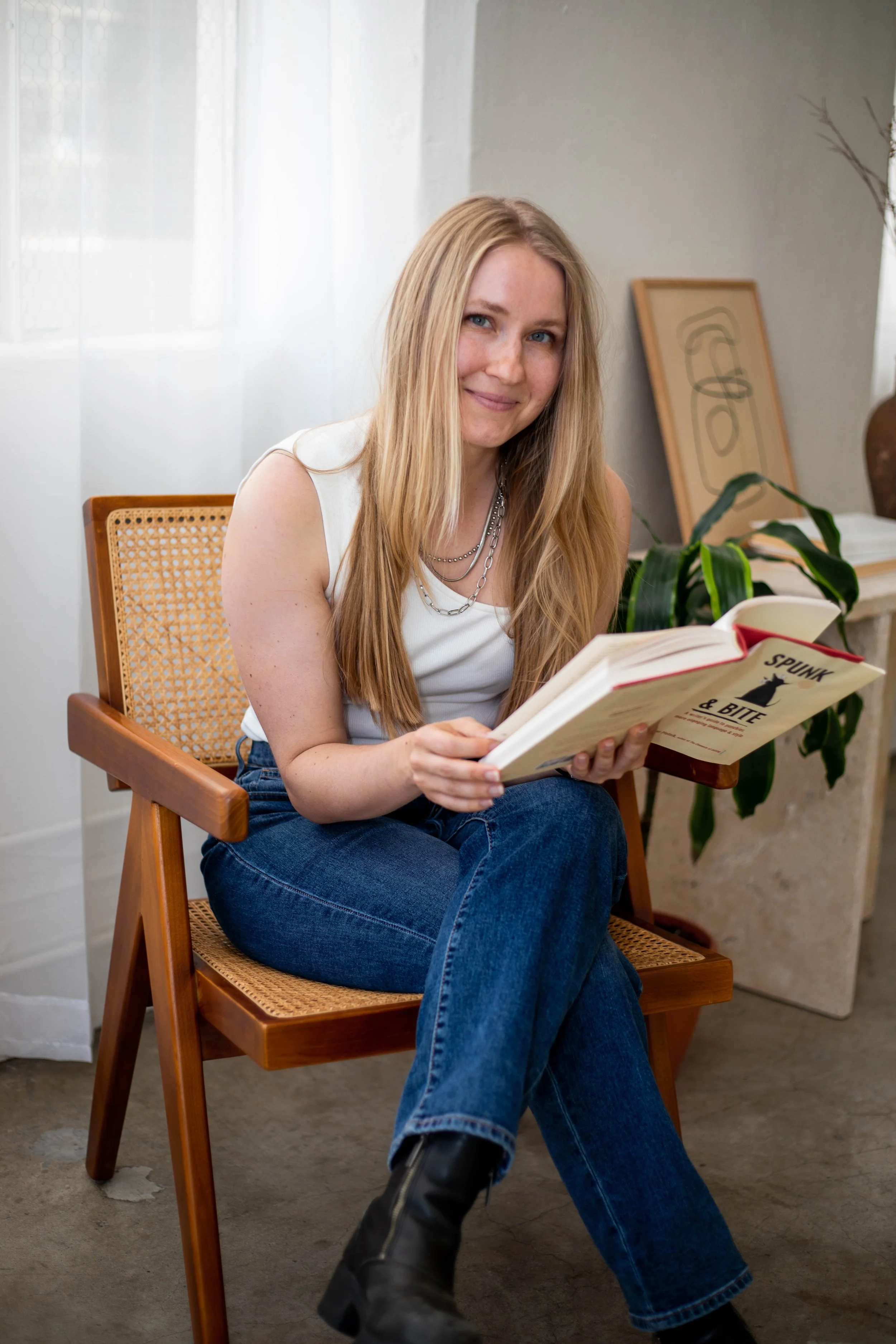 Photo of a white woman with long blonde hair sitting in a chair, holding a book, smiling at the camera