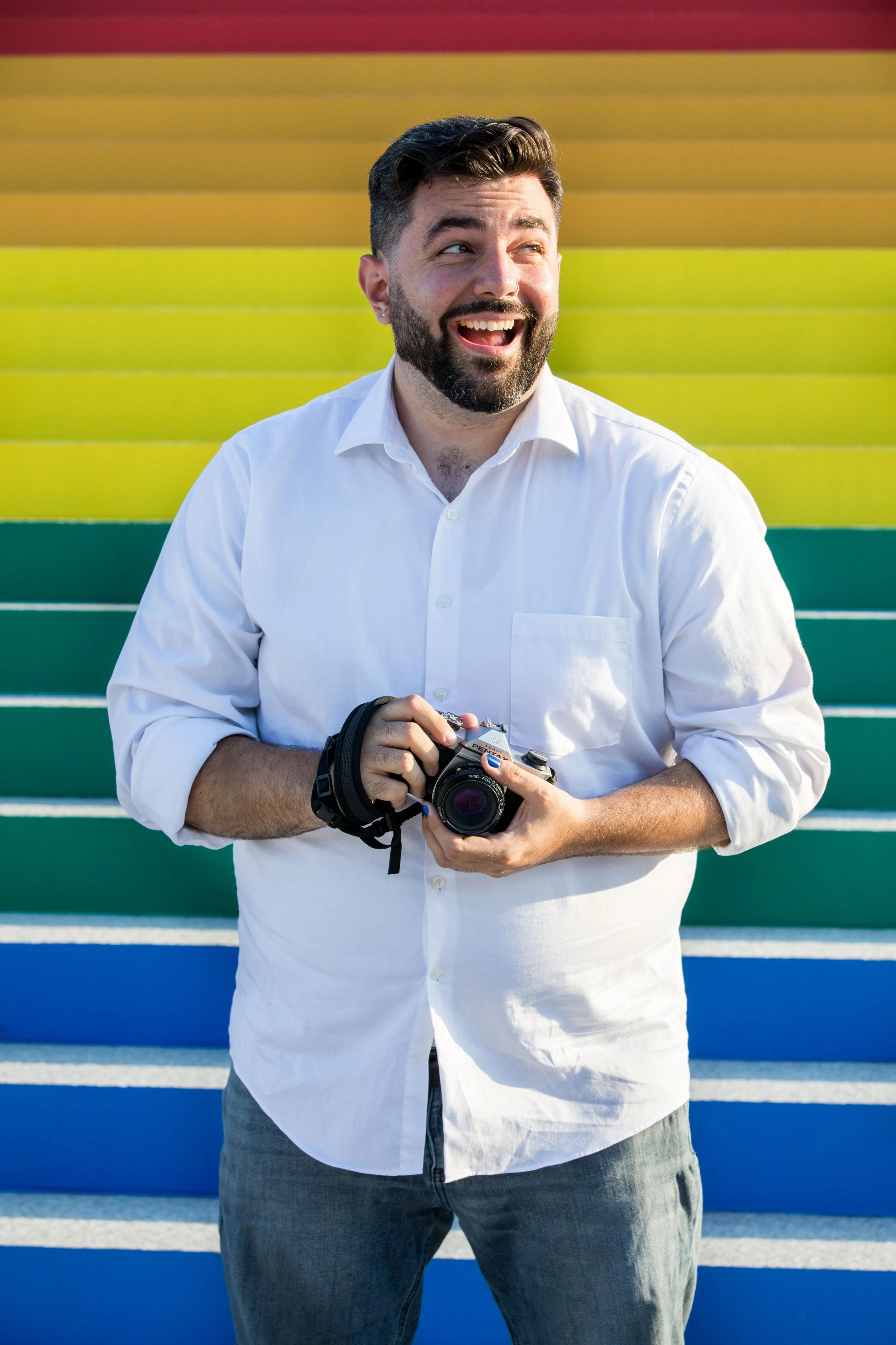 Photo of a white man, standing in front of a rainbow colored wall, holding a camera, smiling