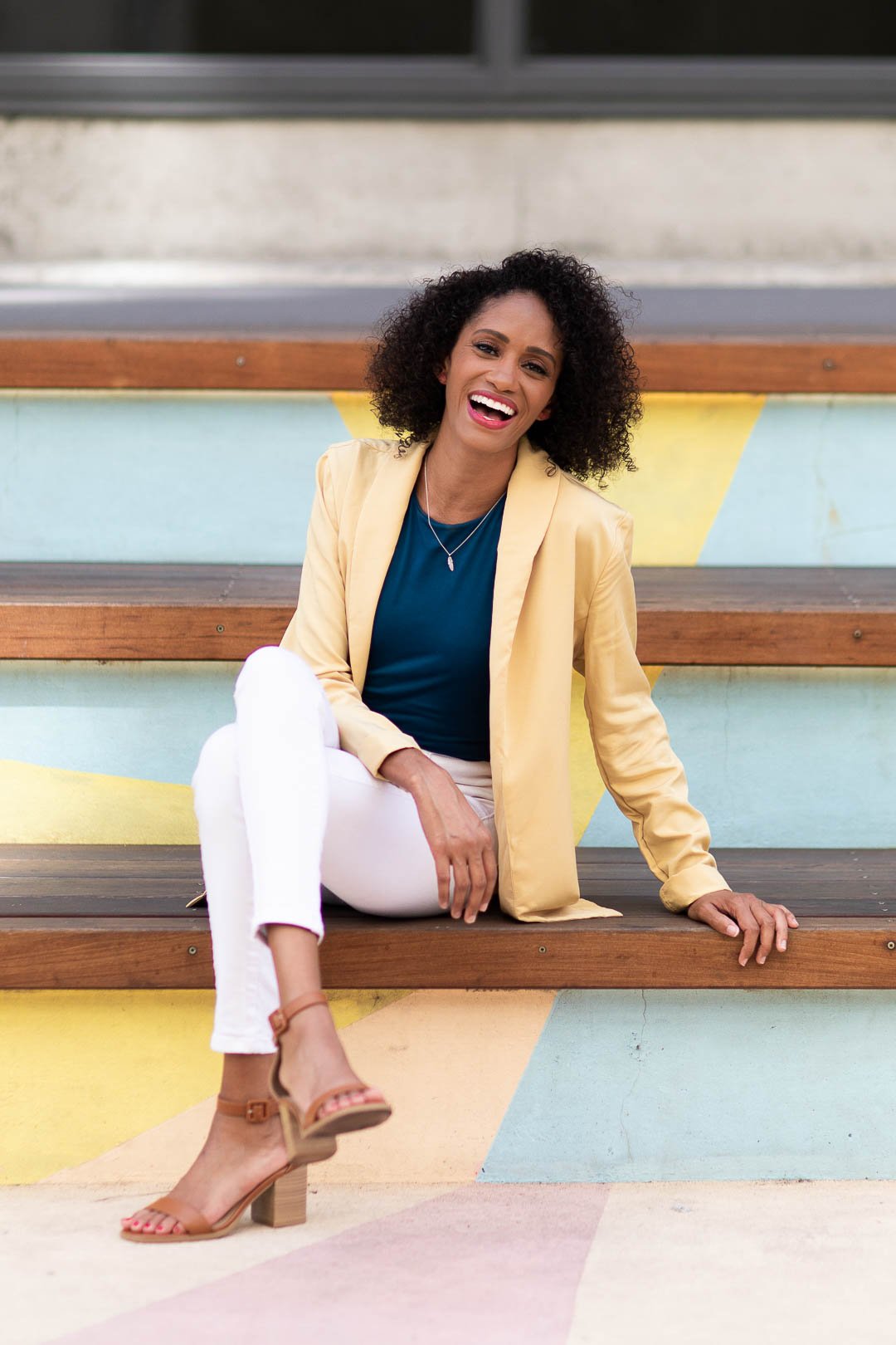 Photo of Crystal, a Black woman with shoulder length dark brown curly hair sitting on a wooden bench, with her legs crossed, smiling at the camera