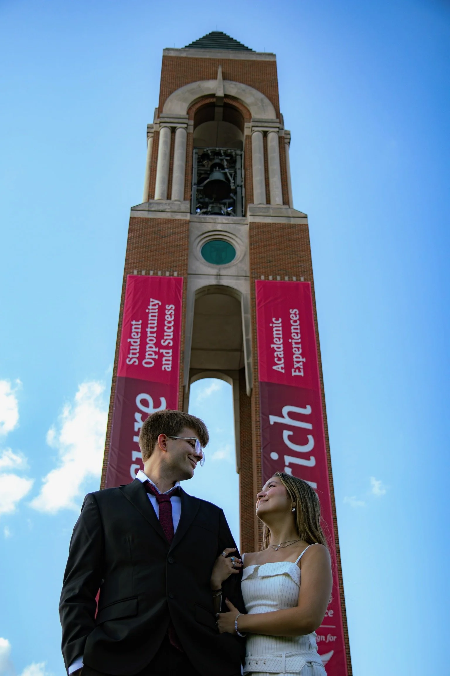 A young man and woman in formal attire stand close and smile at each other outdoors, with a tall brick clock tower with banners in the background against a blue sky with some clouds.