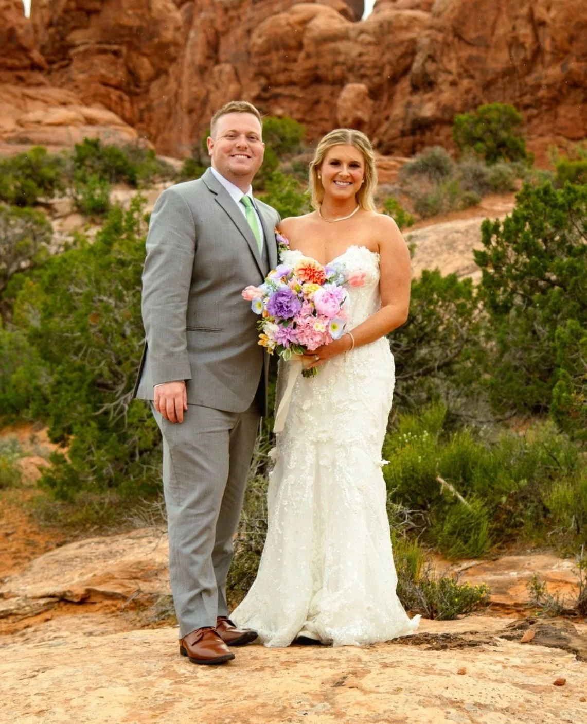 A newlywed couple standing outdoors in a desert landscape, with red rock formations and green shrubs in the background. The groom is wearing a gray suit with a light green tie, and the bride is in a white lace wedding dress holding a colorful bouquet