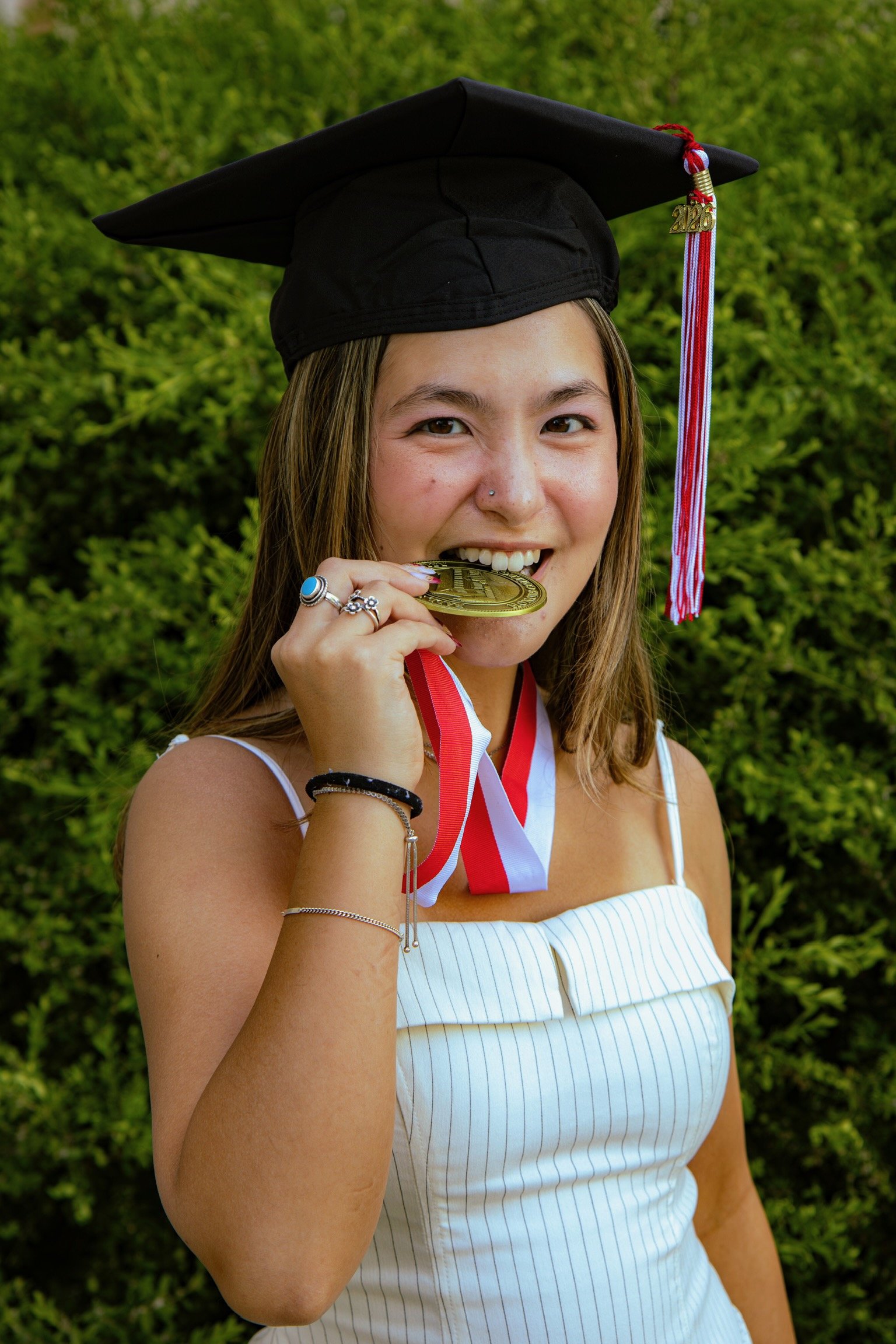 A young woman in a graduation cap and gown biting a gold medal, standing outdoors with green bushes in the background.