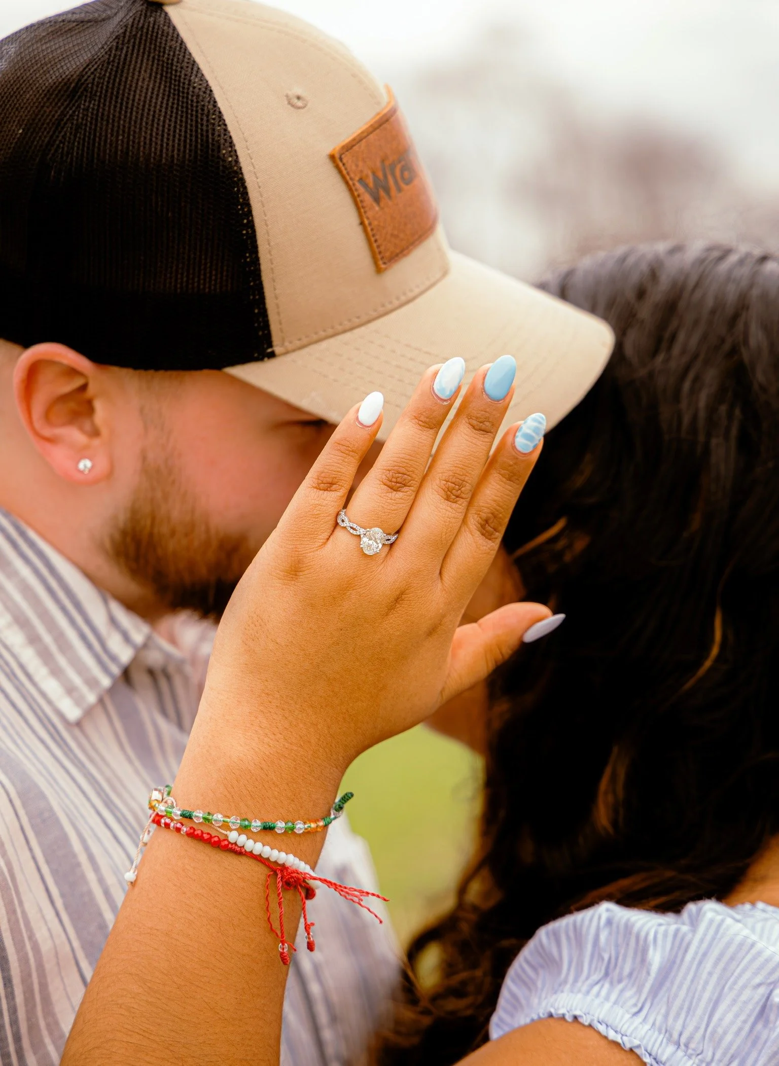 Close-up of a man wearing a beige and black trucker hat. The man has a diamond earring, and colorful bracelets on his wrist. A woman with long dark hair and painted nails rests her head against his. The woman's hand is on the man's forehead with her 