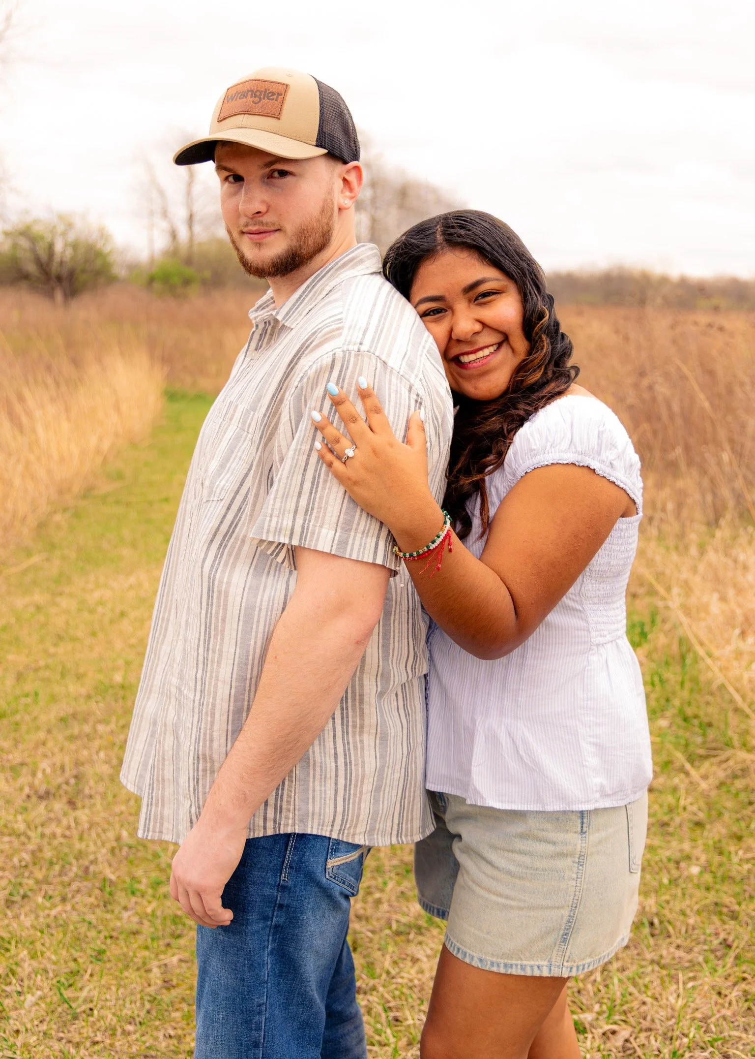 A young man and woman stand back-to-back outdoors in a field with dry grass and sparse trees, smiling and looking at the camera. The man wears a beige and black trucker hat and a light striped shirt, while the woman wears a white top and shorts with 