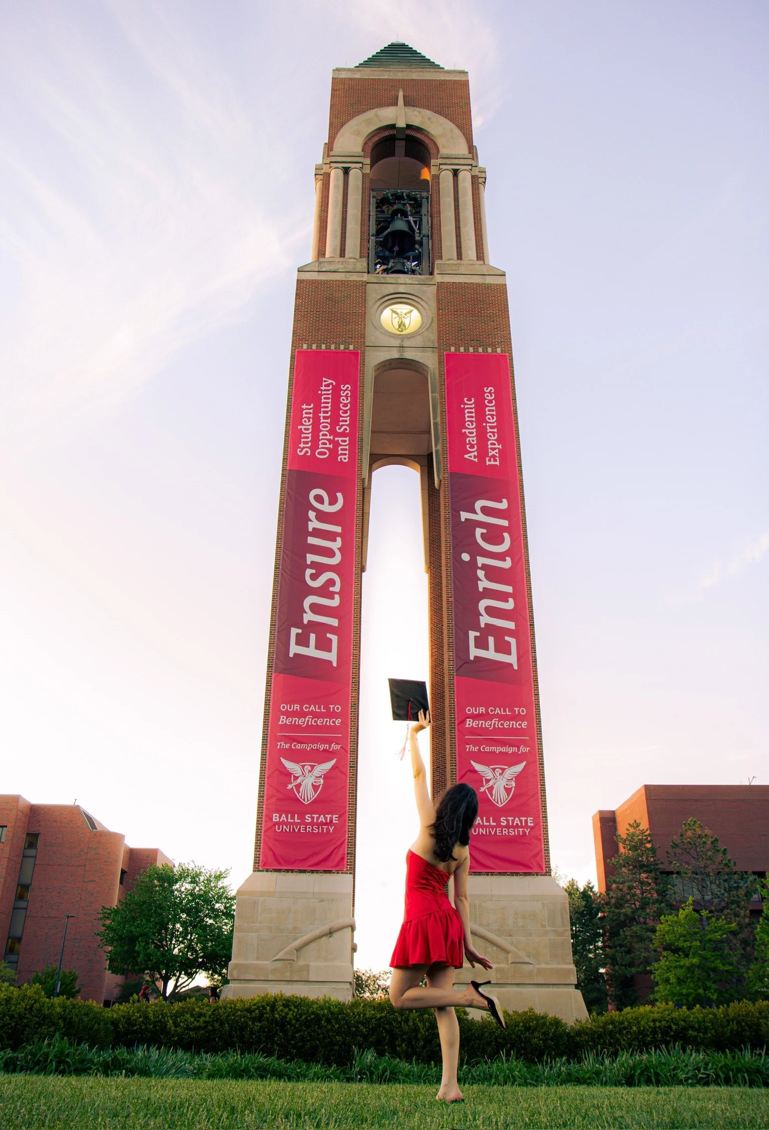 A woman in a red dress and high heels performs a dance pose with one knee bent and one arm raised in front of a university clock tower with banners that read 'Enrich', 'Student Opportunity and Success', 'Academic Experiences', and 'Ball State Univers