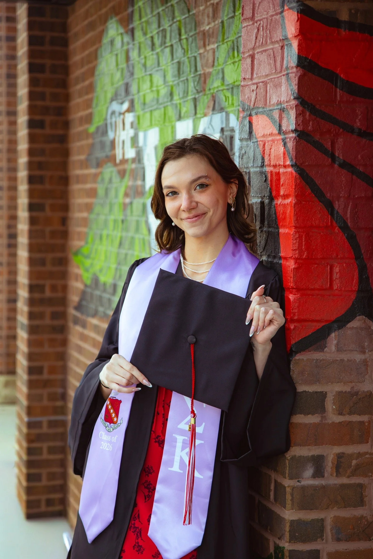 A young woman in graduation attire holding her graduation cap, standing outdoors against a brick wall decorated with colorful graffiti art. She is smiling and has earrings and an eyebrow piercing.