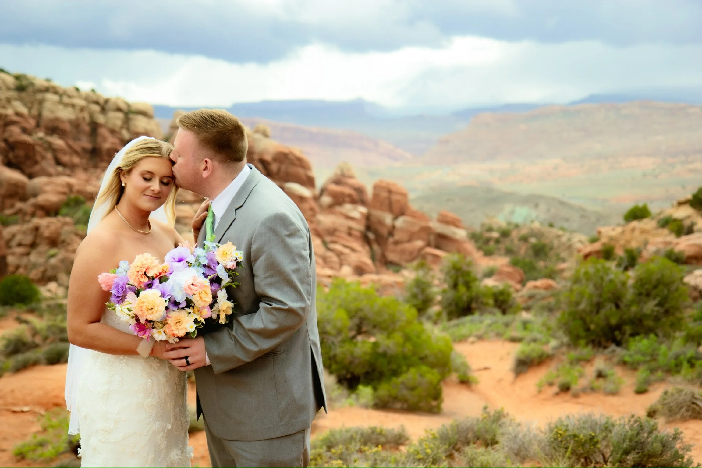 A bride and groom on their wedding day, standing close together outdoors in a desert-like landscape with red rock formations and a cloudy sky in the background. The bride is holding a large bouquet of colorful flowers, and they are sharing an intimat