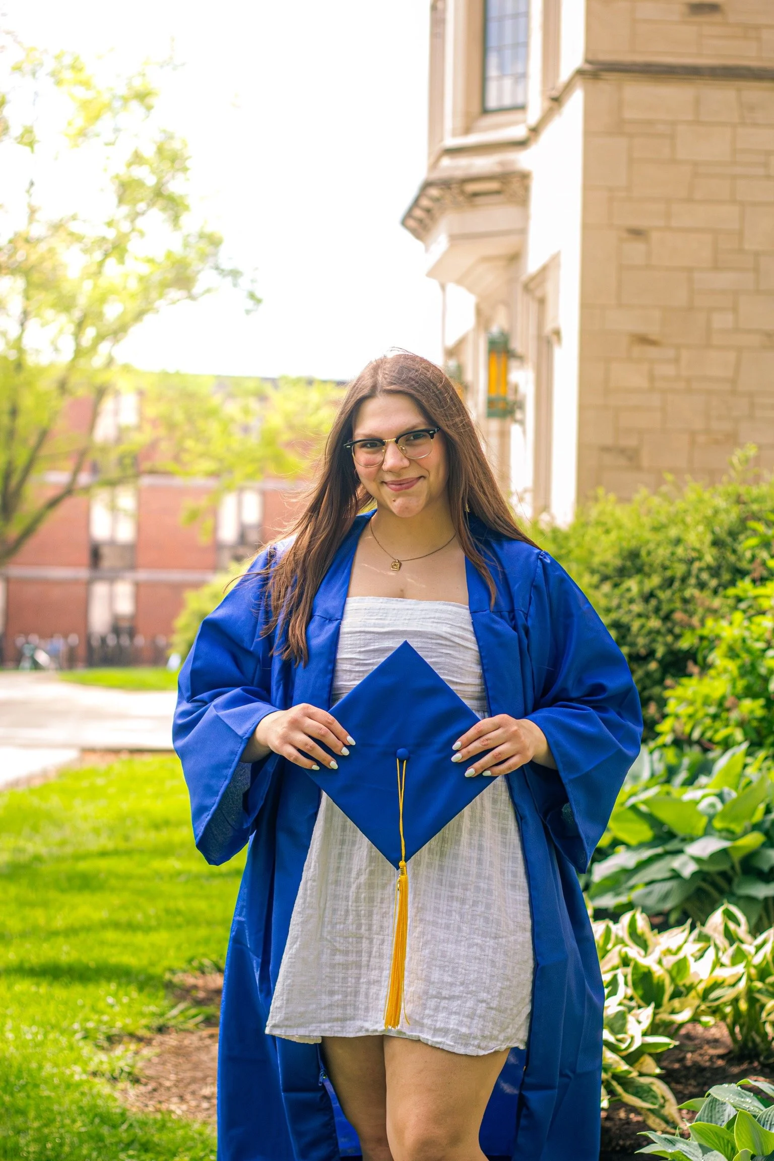 Young woman in a blue graduation gown holding a matching mortarboard hat, standing outdoors on a garden path with trees and a brick building in the background.