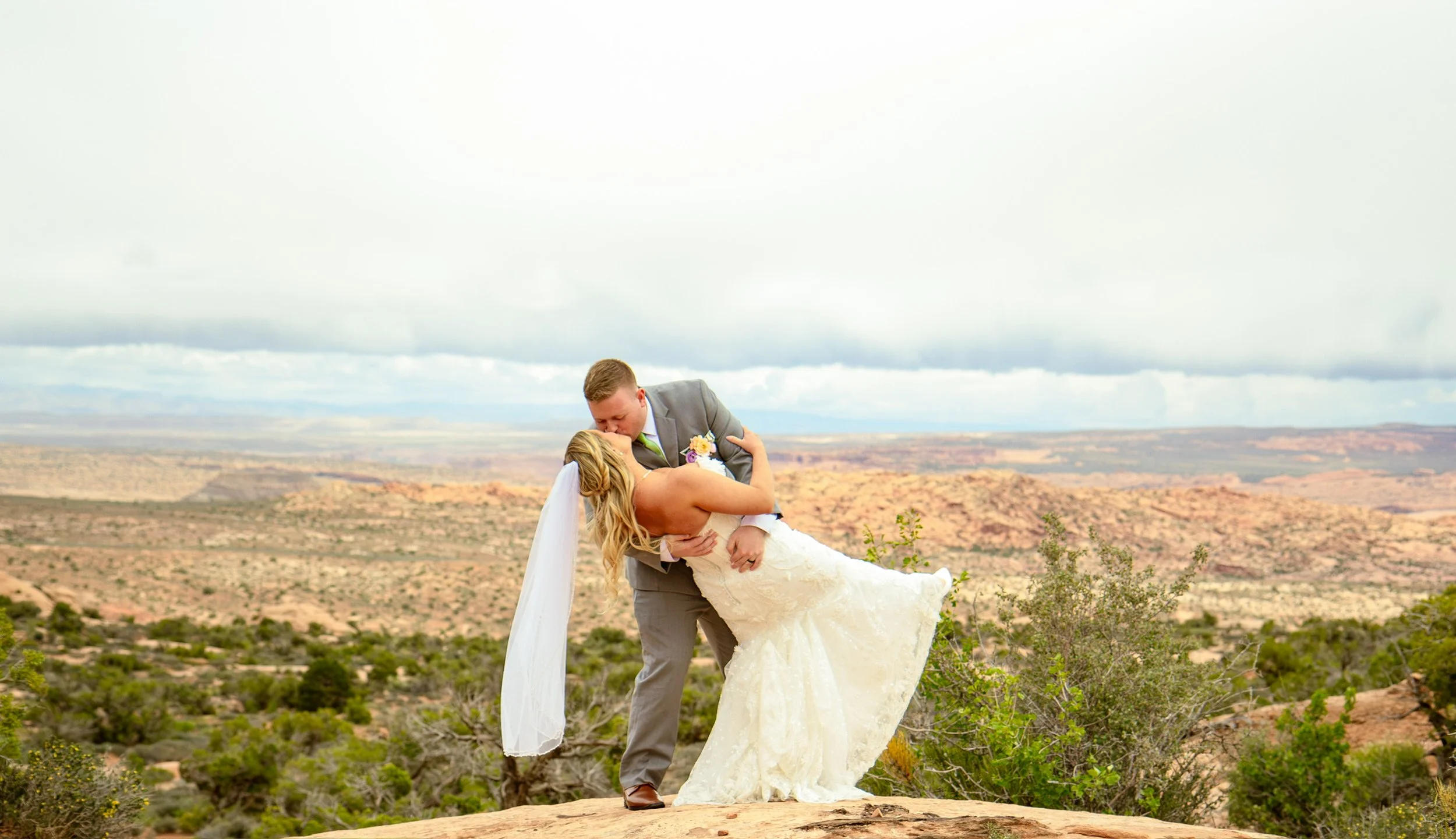 A groom lifting a bride in a desert landscape with rocky terrain and sparse green vegetation, under a cloudy sky.