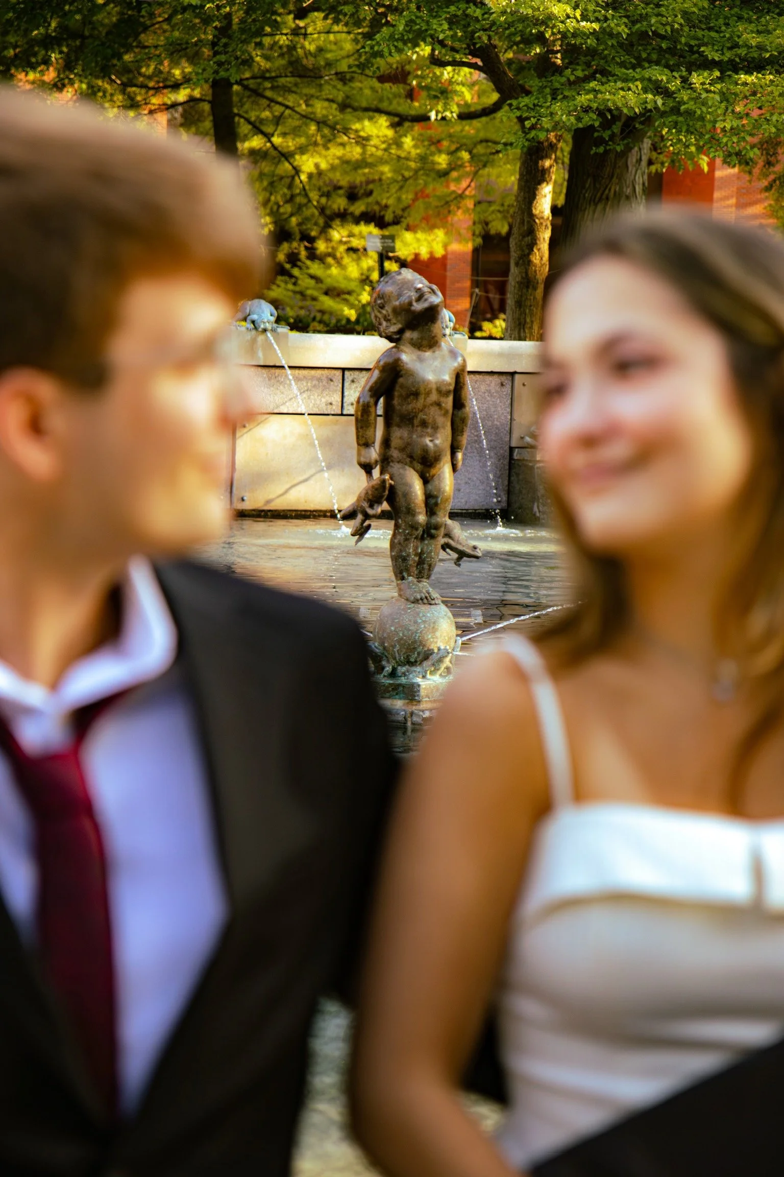 Two people standing in front of a fountain with a bronze statue of a young boy, surrounded by trees with green leaves in a park or garden setting.