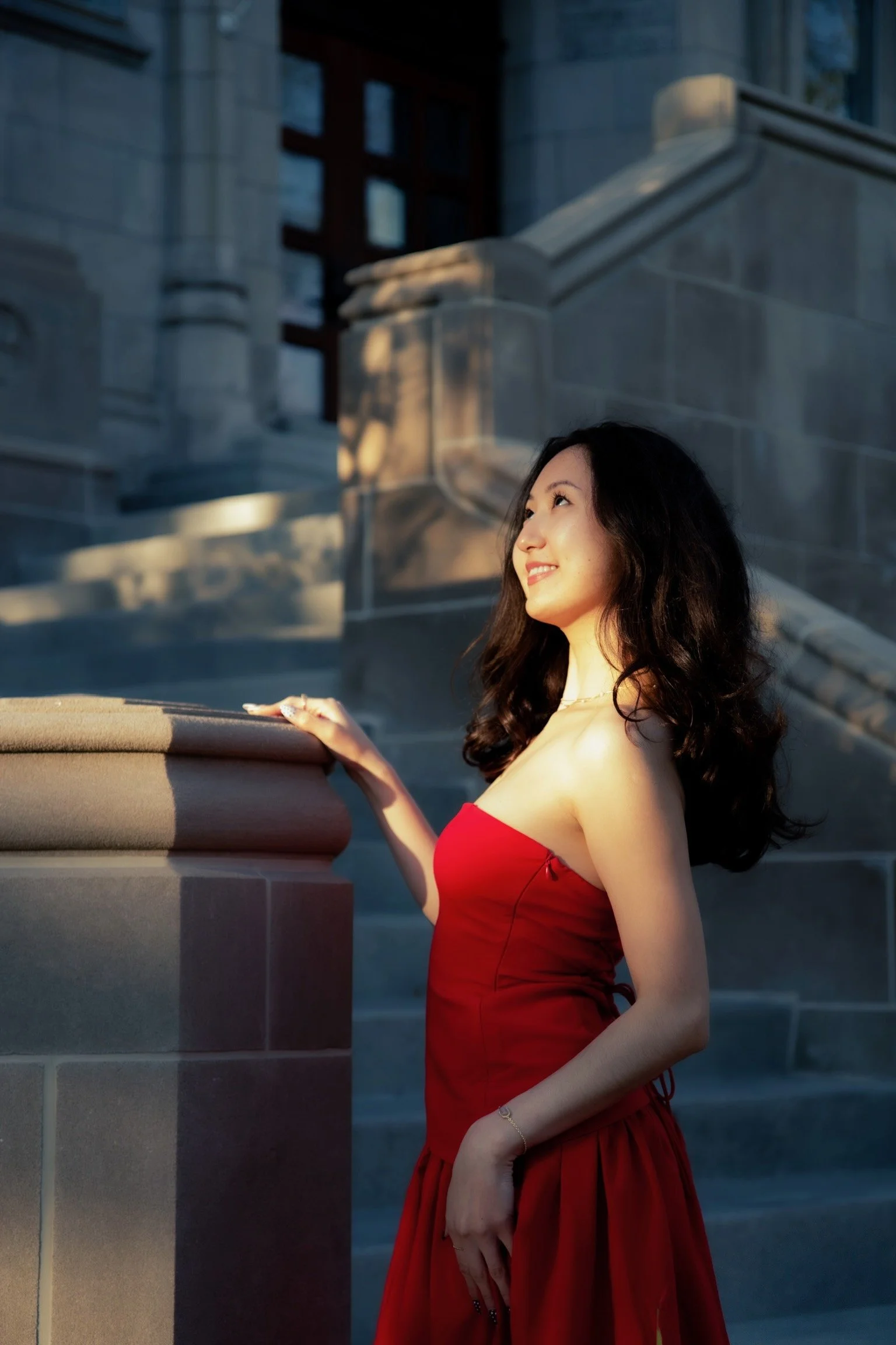 A young woman with long dark wavy hair wearing a strapless red dress standing outdoors at a historic building with stone stairs and columns during sunset, smiling and looking to her left.