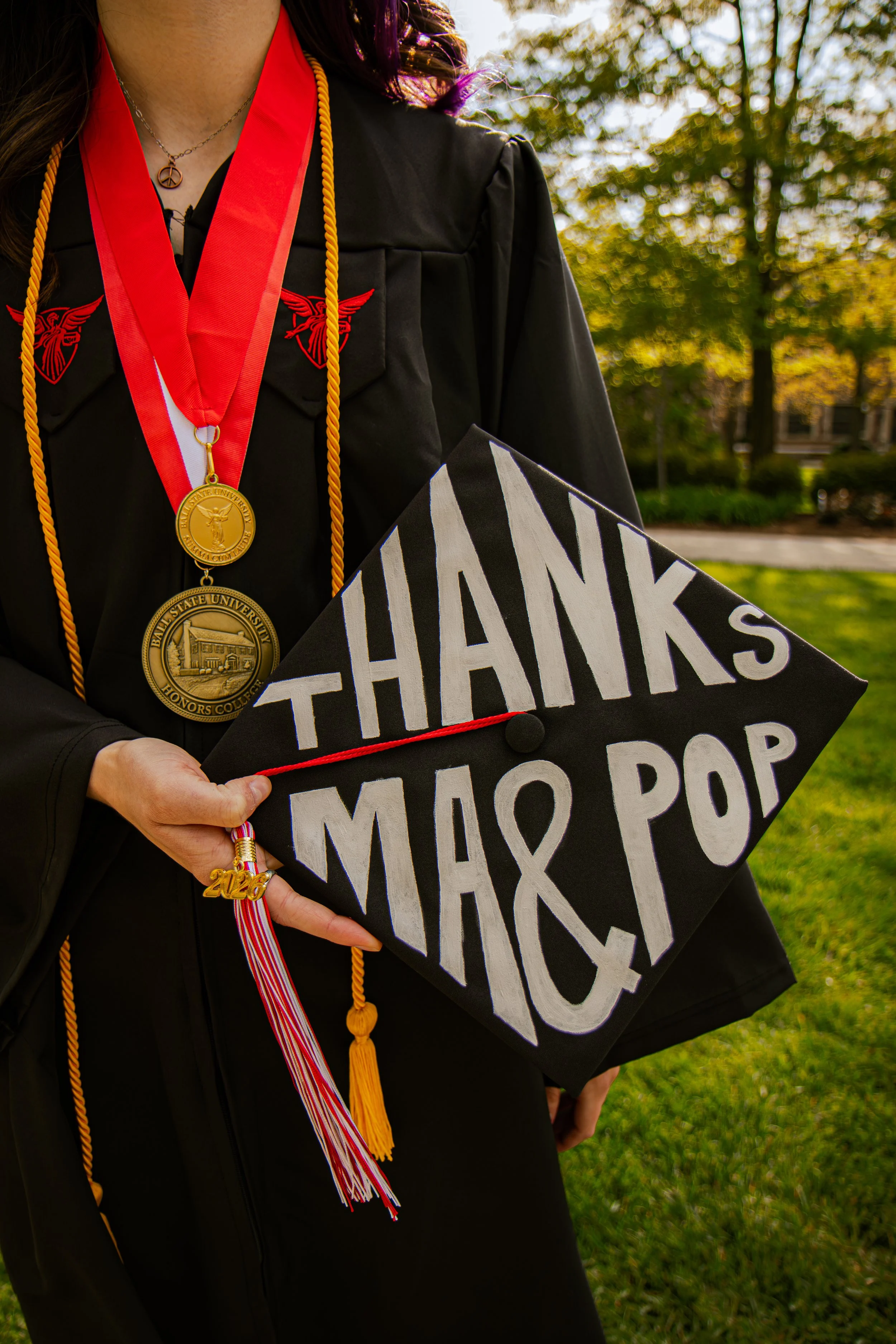 A person in a black graduation gown with red and yellow cords and medals. The person is holding a cap that says 'THANKS MIO POP' with a background of trees and grass, likely during a graduation ceremony.
