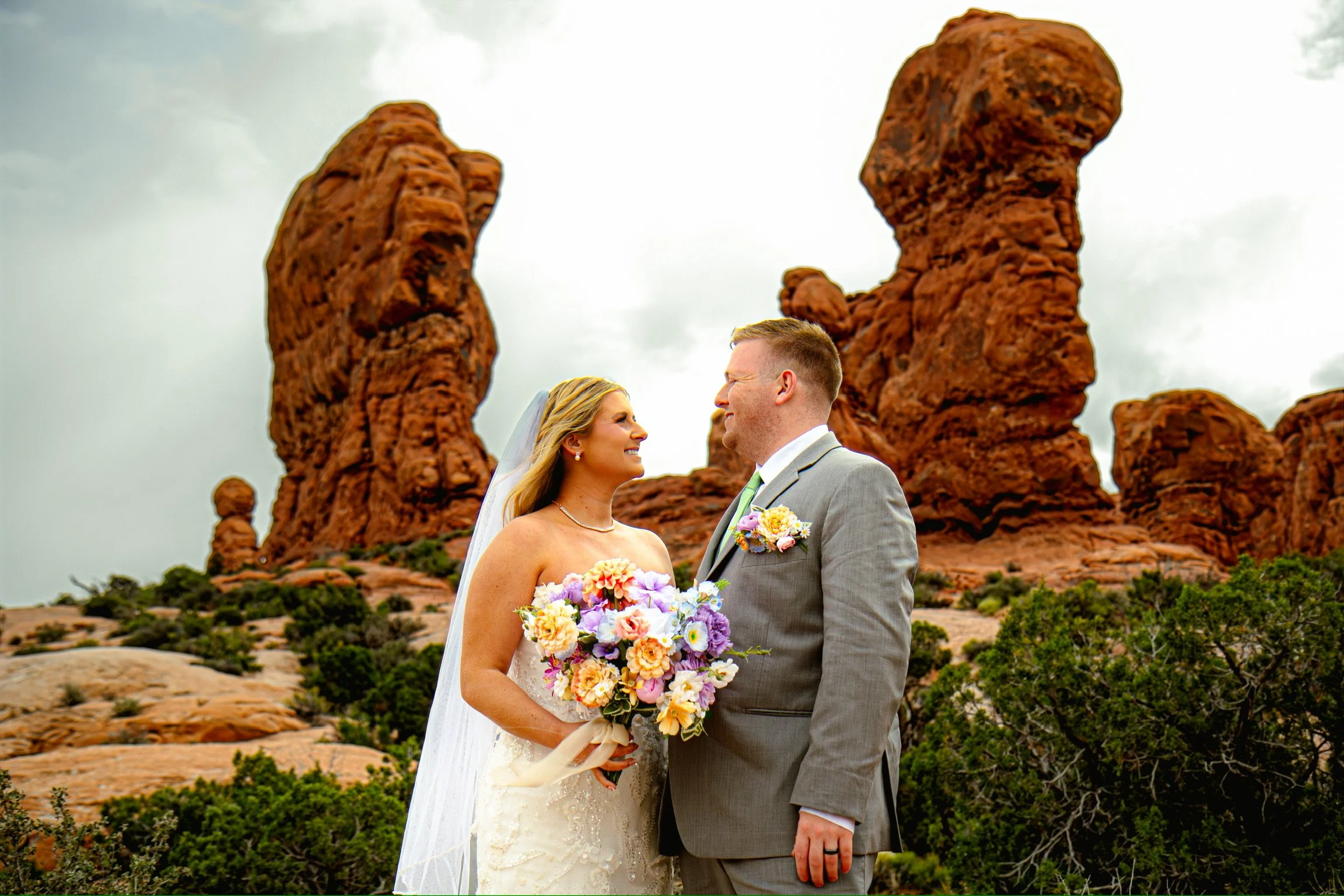 A bride and groom standing face to face outdoors during a wedding ceremony, with red rock formations and green vegetation in the background.