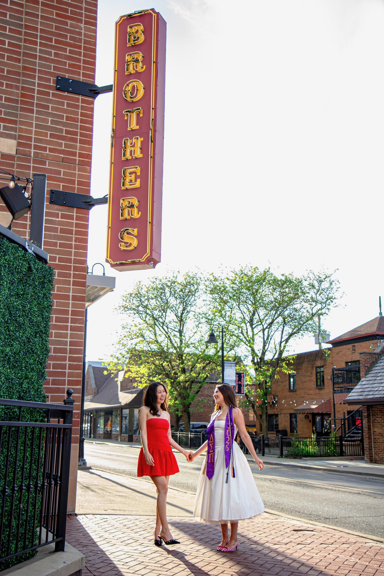 Two women holding hands and smiling on a city street, with a sign for a bar above them and trees and buildings in the background.
