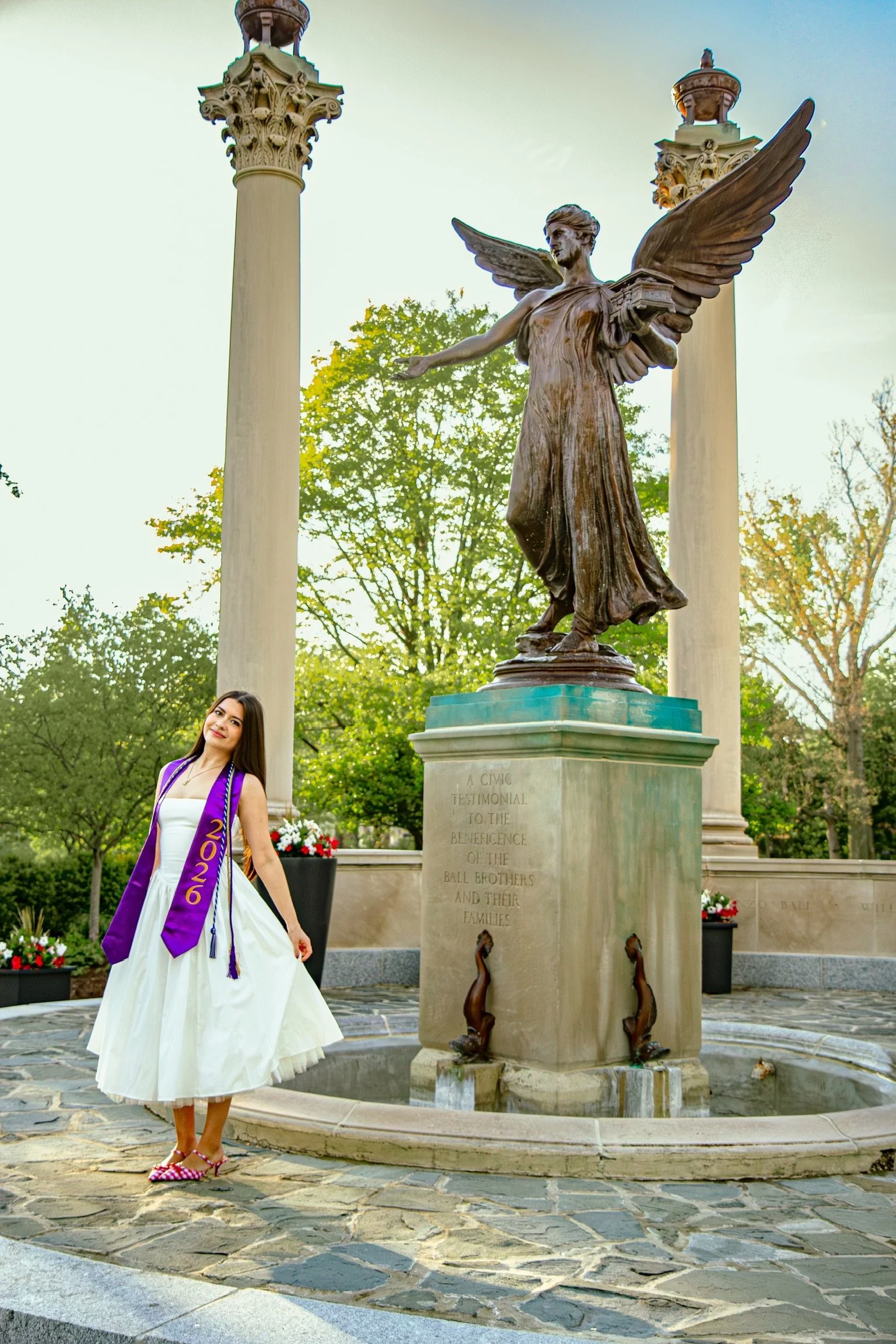 A young woman in a white dress and pink polka dot shoes wearing a purple sash with gold writing stands next to a bronze statue of a winged figure holding a book, in a park with trees, flowers, and tall white pillars in the background.