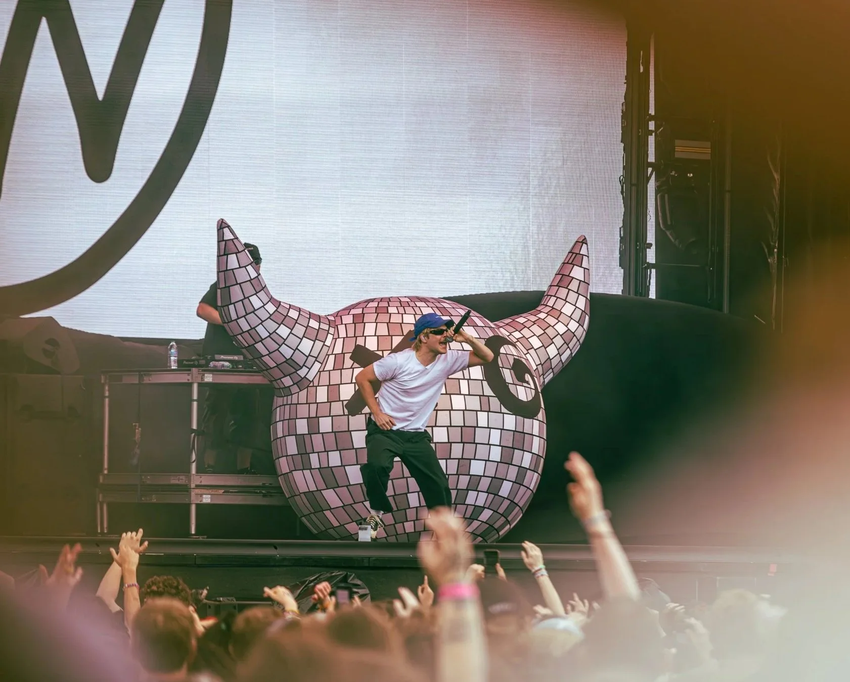 A male performer wearing a white t-shirt, black pants, a blue cap, and sunglasses singing into a microphone on stage at a music festival with a colorful abstract sculpture of a pig's head in the background and hands raised in the audience.