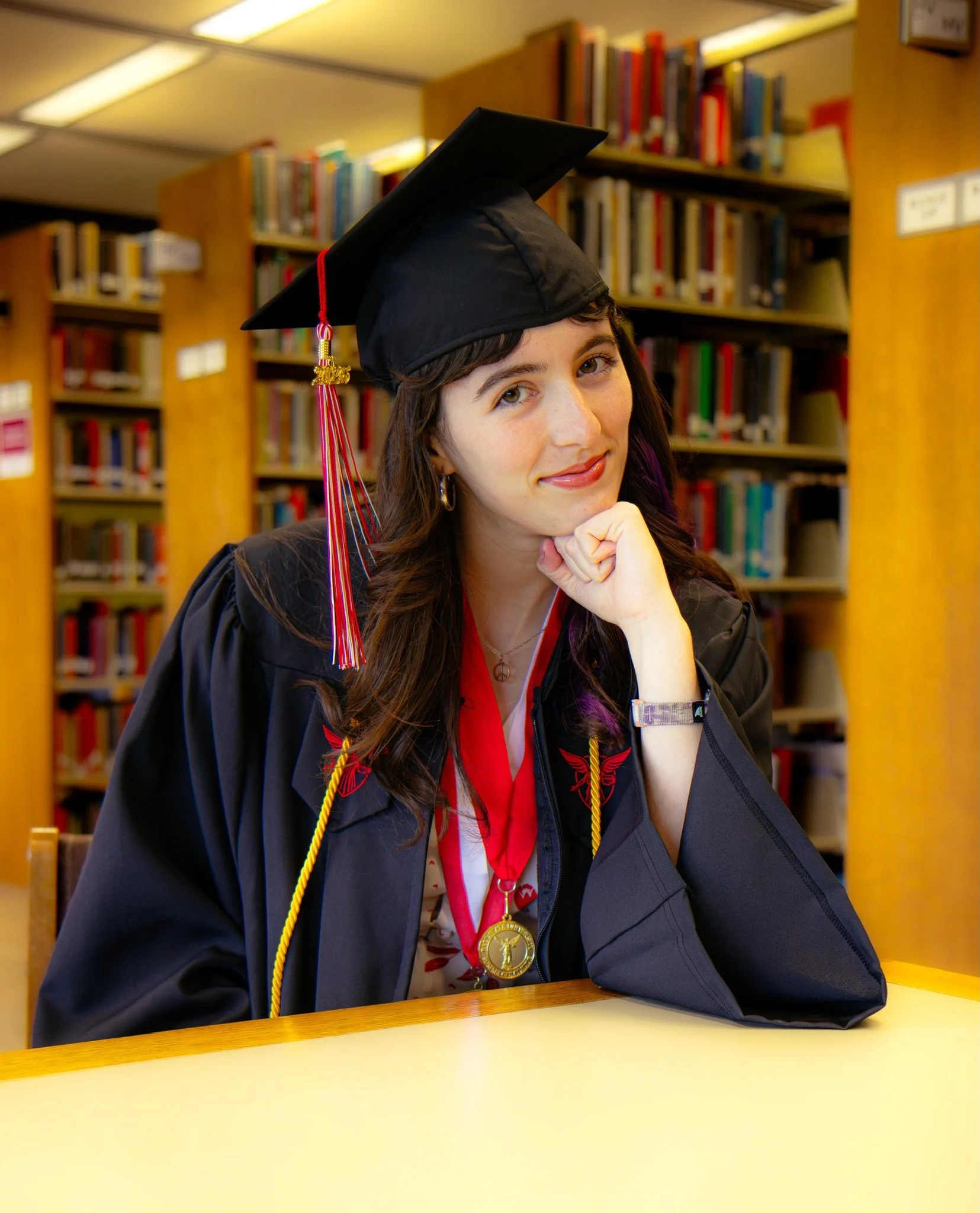 A young woman in a graduation cap and gown sitting at a table in a library, wearing a medal around her neck, with bookshelves in the background.