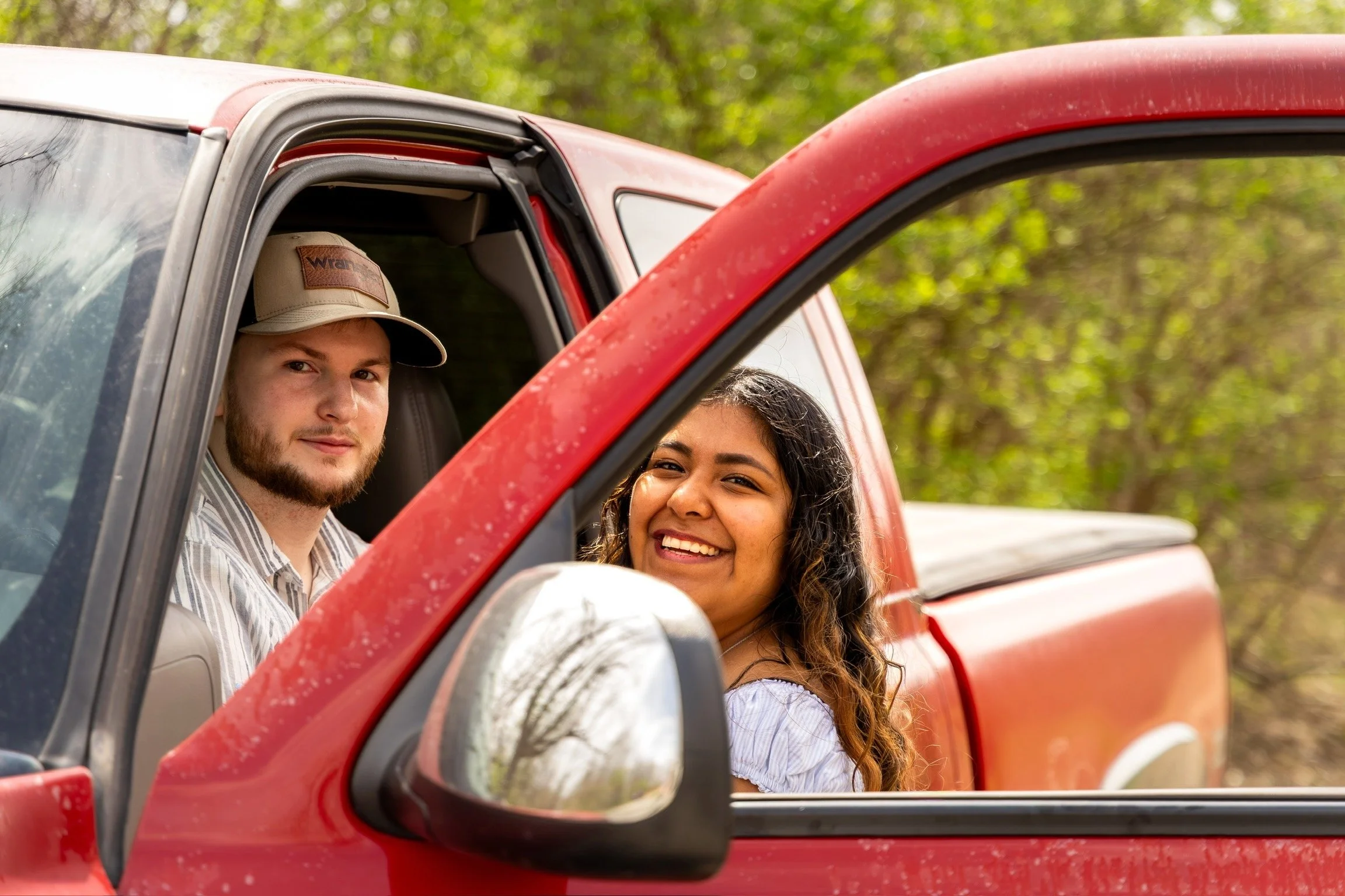 A man and woman sitting in a red pickup truck, smiling and looking out of the open window. The background features a lush green, wooded area.