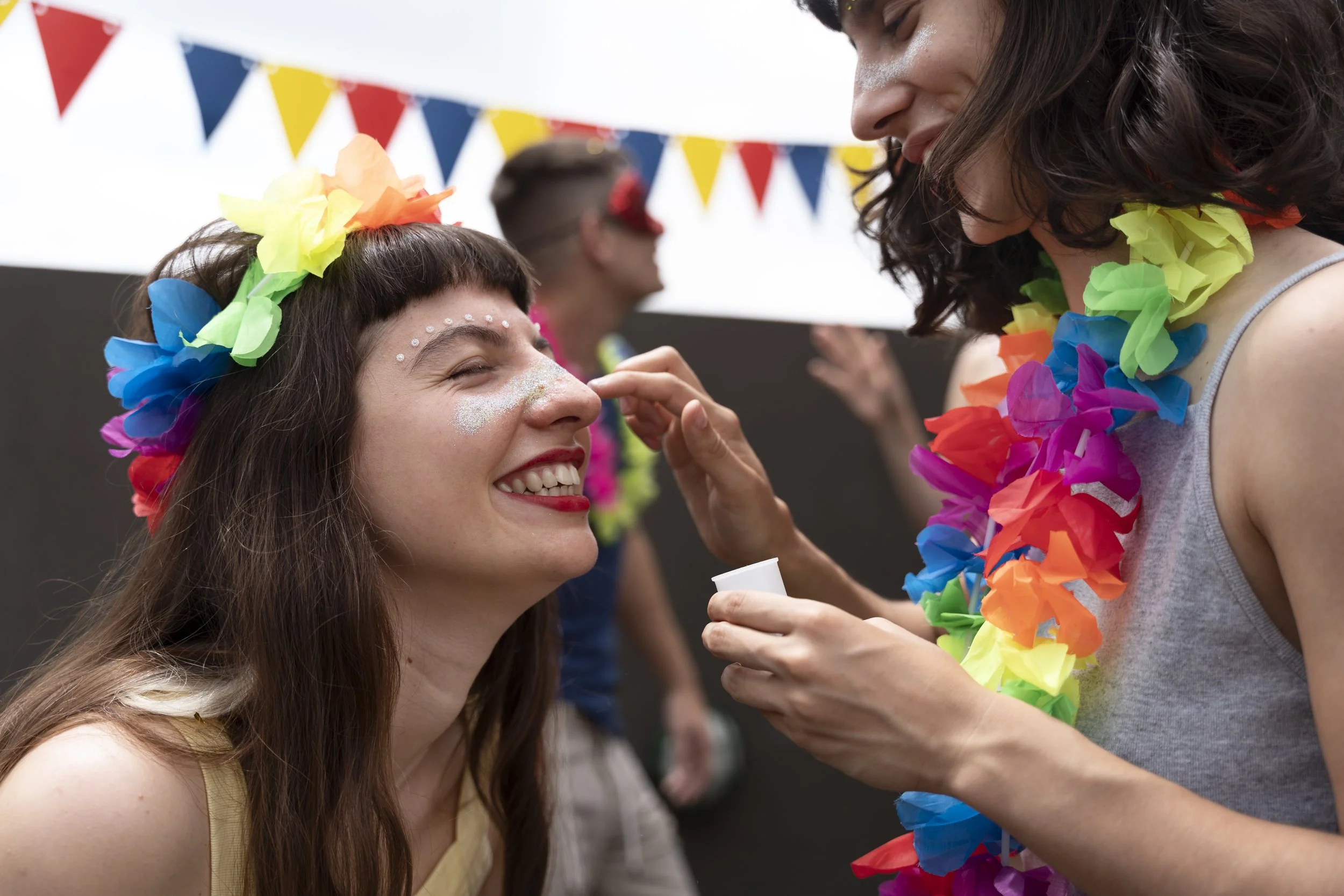 Women celebrating at a festive event, one woman is smiling with a flower crown on her head while another is applying makeup, wearing a colorful lei, against a background of colorful bunting.