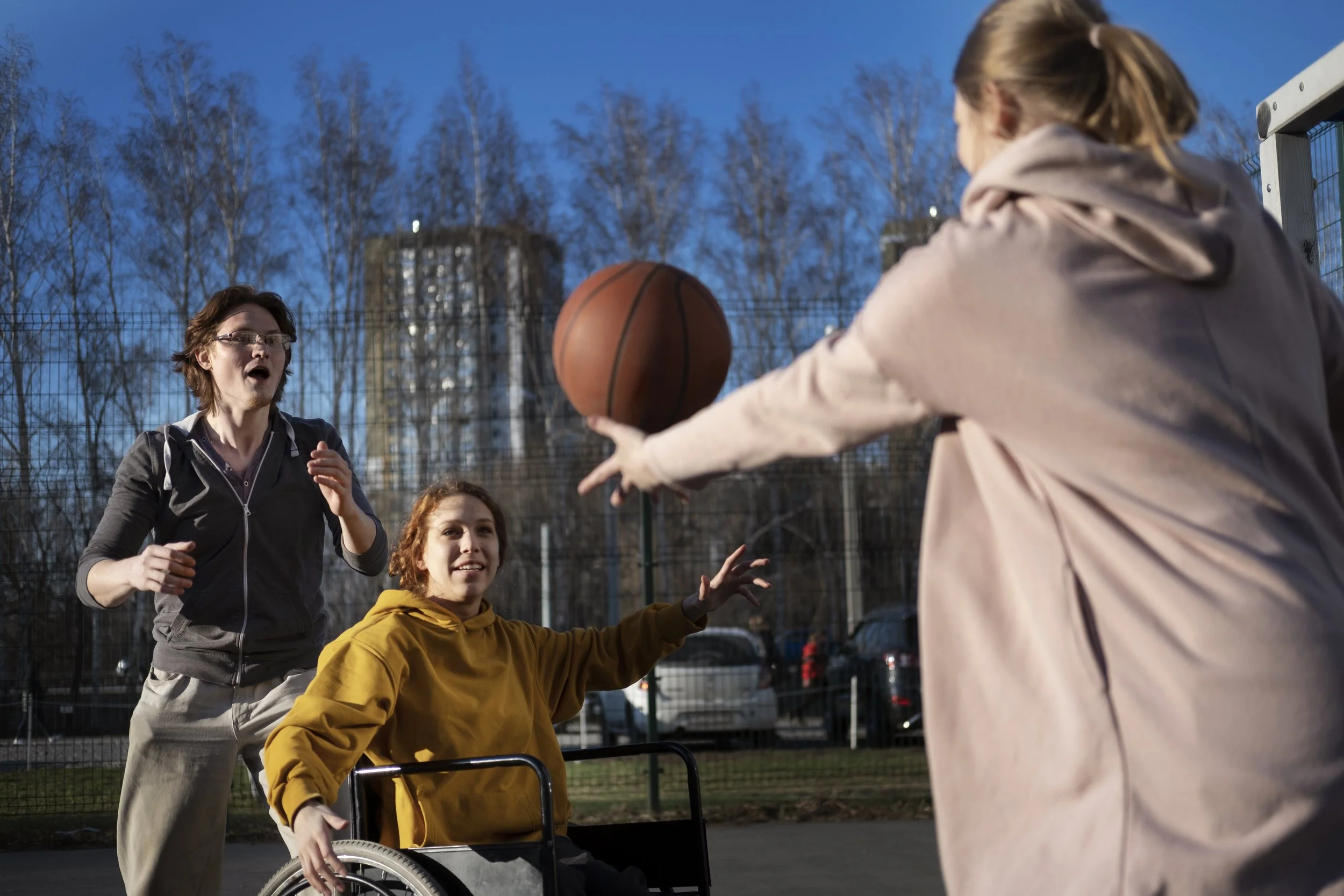 Three women playing wheelchair basketball outdoors in the evening, with trees and buildings in the background.