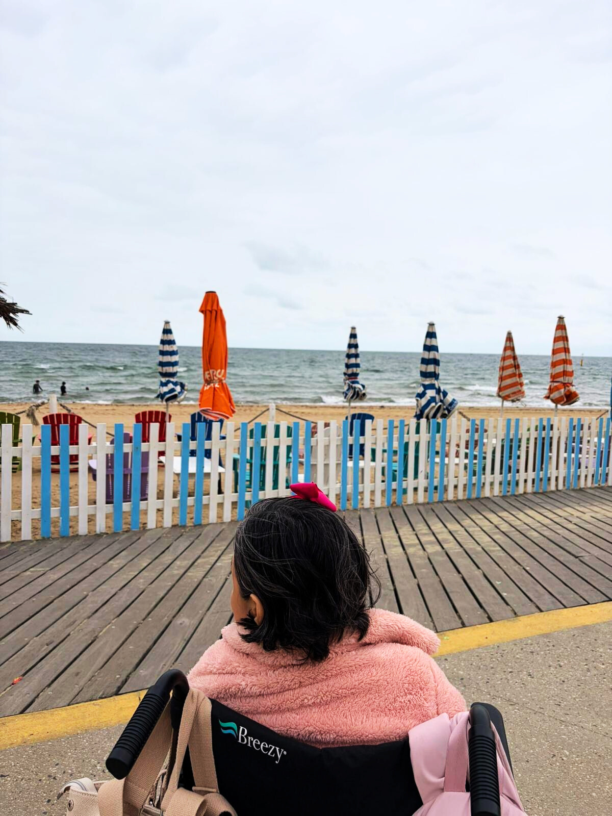 A woman in a pink jacket sitting in a wheelchair on a boardwalk near a beach with striped umbrellas and ocean in the background.
