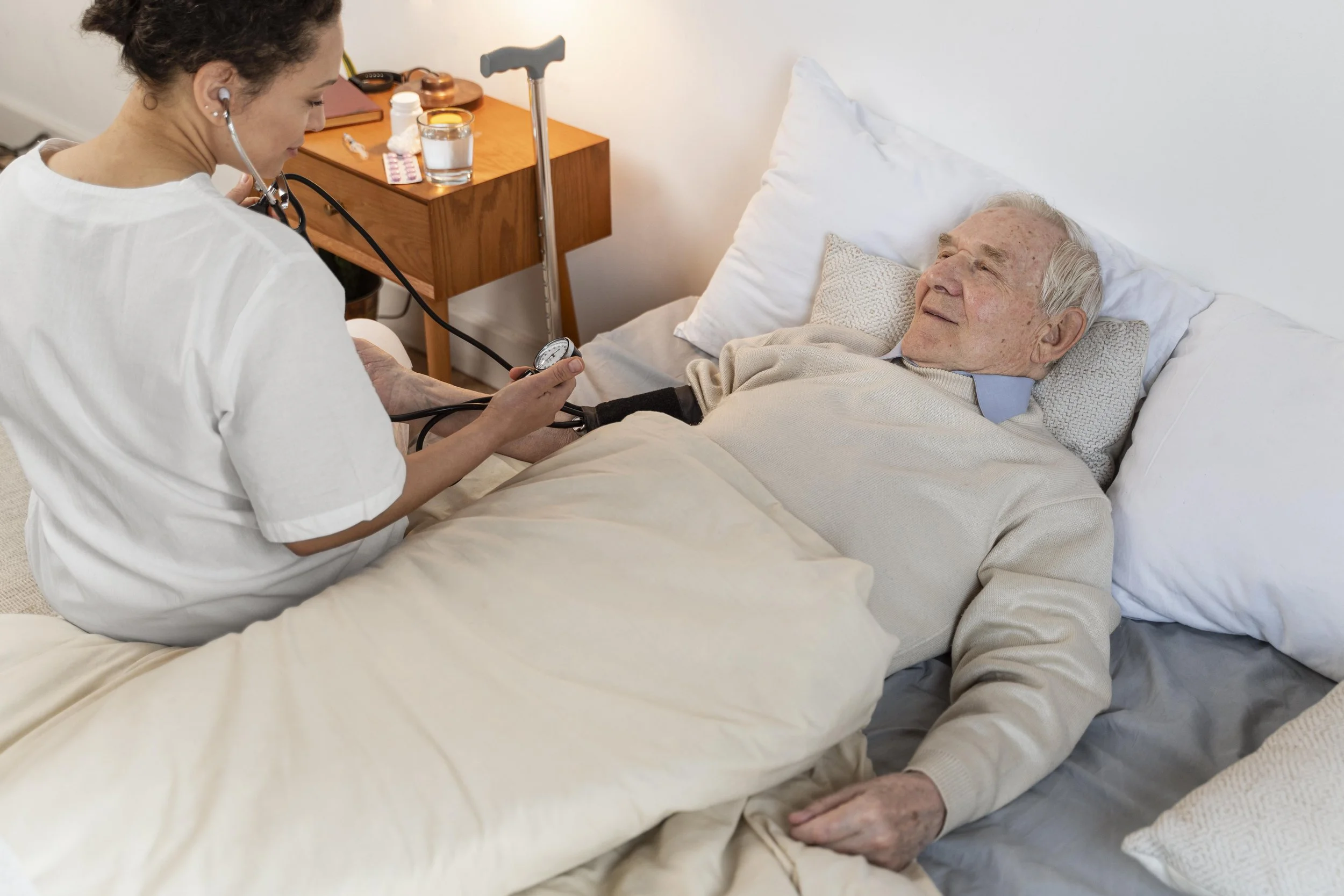 A healthcare worker using a stethoscope to check an elderly man's blood pressure while he lies in bed at home.