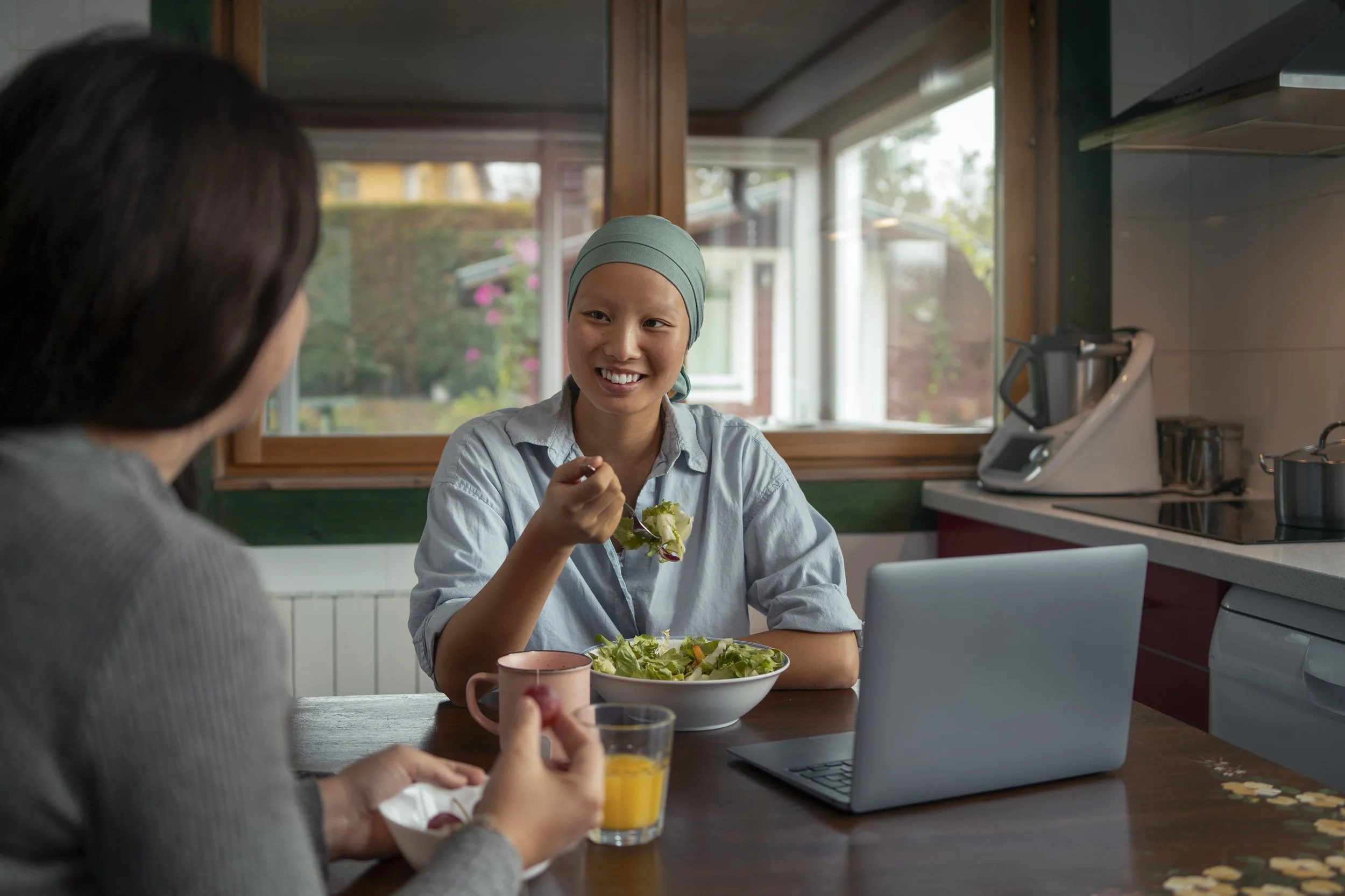 A woman with a headscarf eating a salad and smiling at another woman with dark hair in a kitchen, with a laptop, a glass of orange juice, and a pink mug on the table.