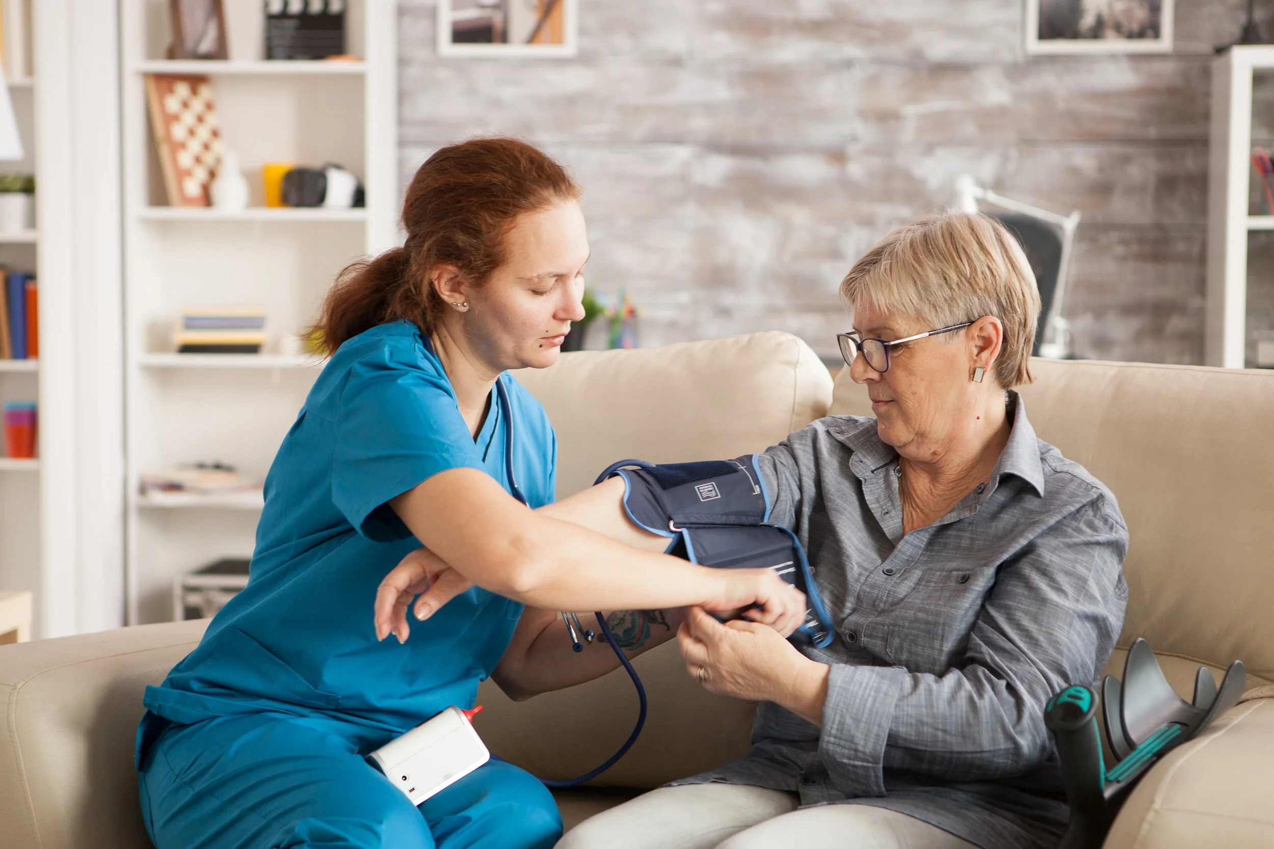 A nurse or caregiver taking an elderly woman's blood pressure at home, sitting on a beige couch in a living room with shelves and picture frames.