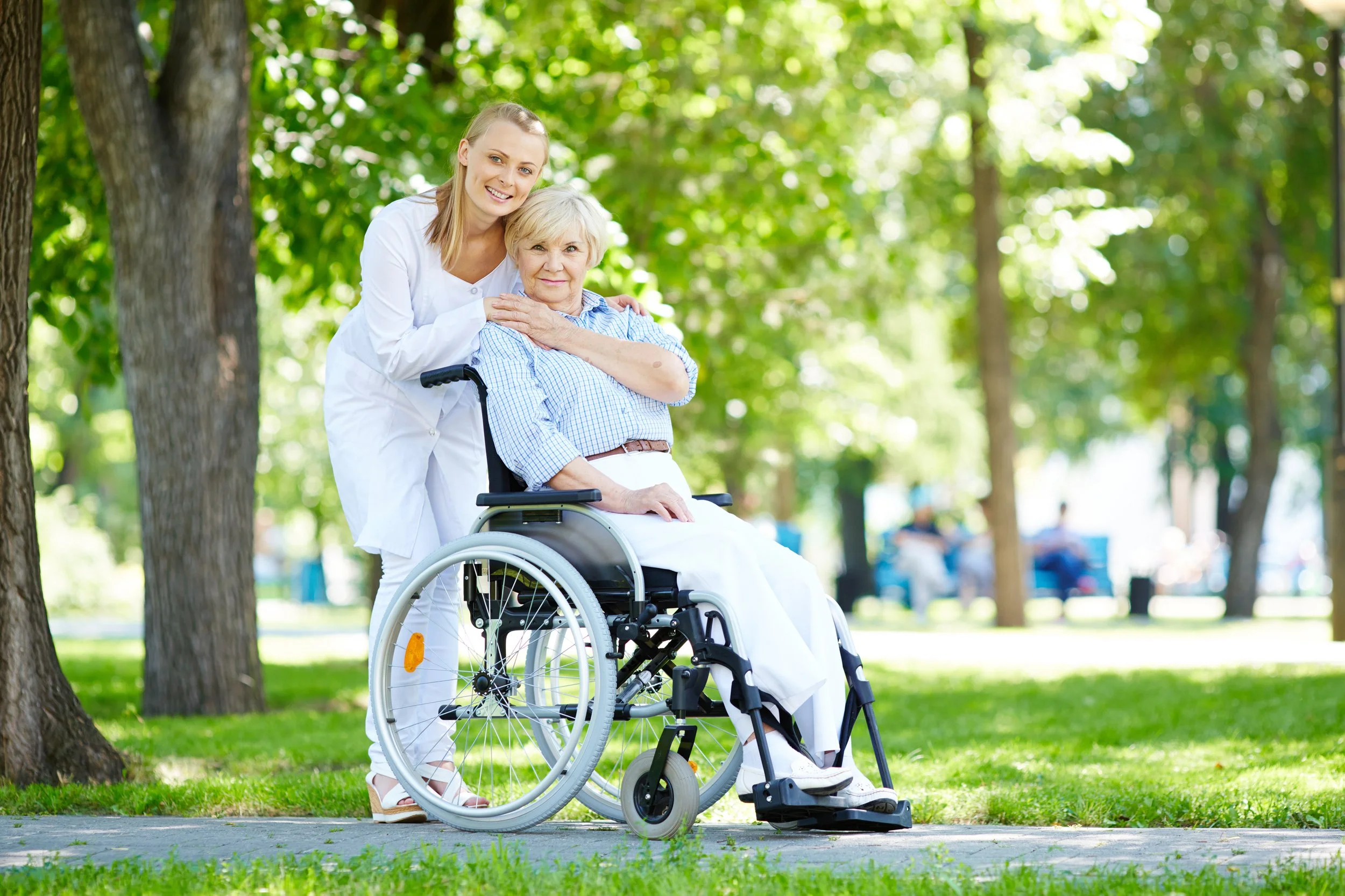 A caregiver woman standing beside an elderly woman in a wheelchair, outdoors in a park with green trees, smiling at the camera.
