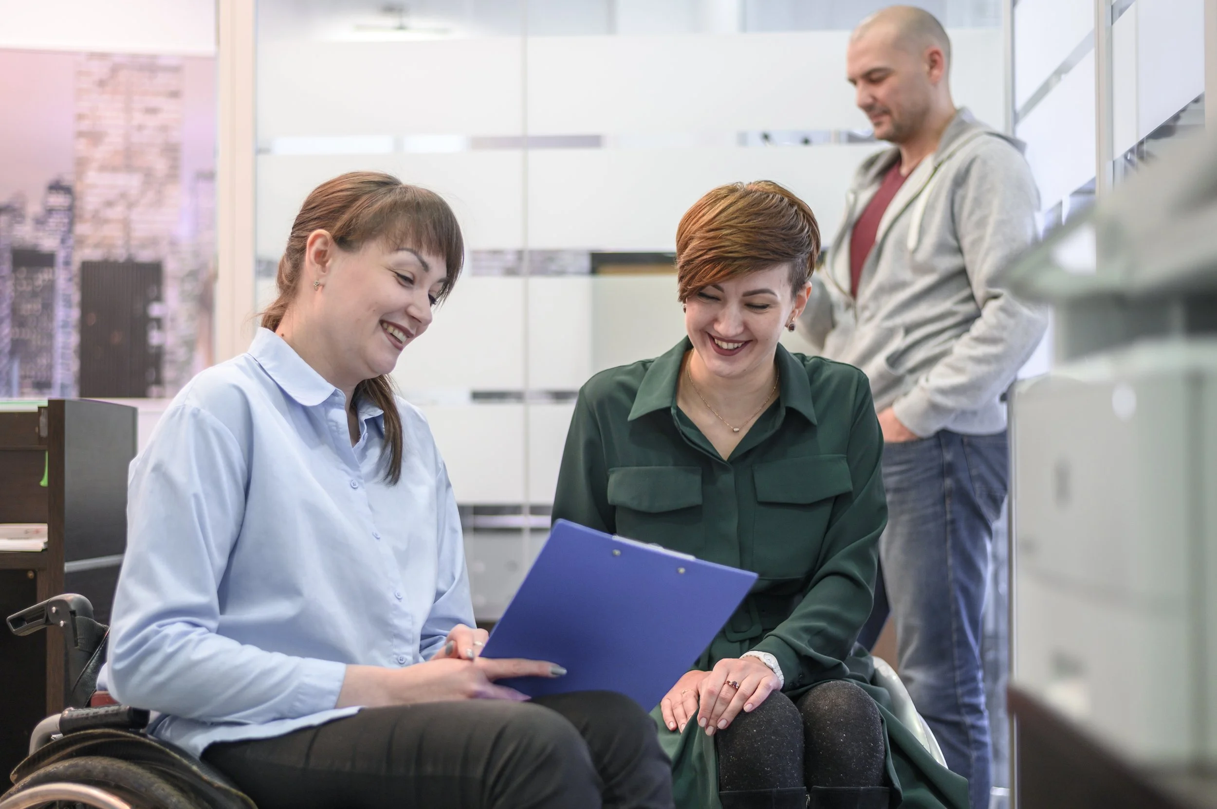 Two women and one man in an office, with the women looking at a document or clipboard, smiling, while the man stands behind them looking on, in a modern office space.