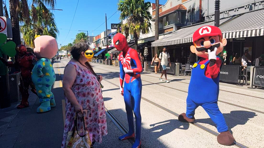 A woman with an emoji face attends a street event with people dressed as Spider-Man, Mario, and a character in a colorful costume. The scene is set on a sunny street lined with shops and palm trees.