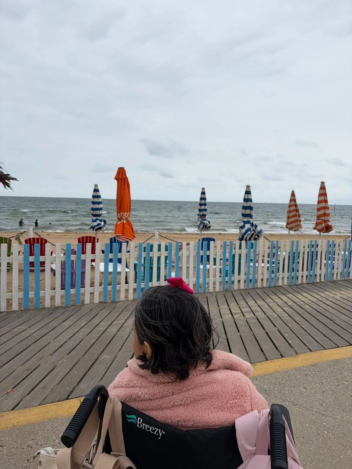 A woman with dark hair in a pink jacket sitting in a Breezy wheelchair, overlooking a beach with closed striped blue and red umbrellas and a colorful wooden fence.
