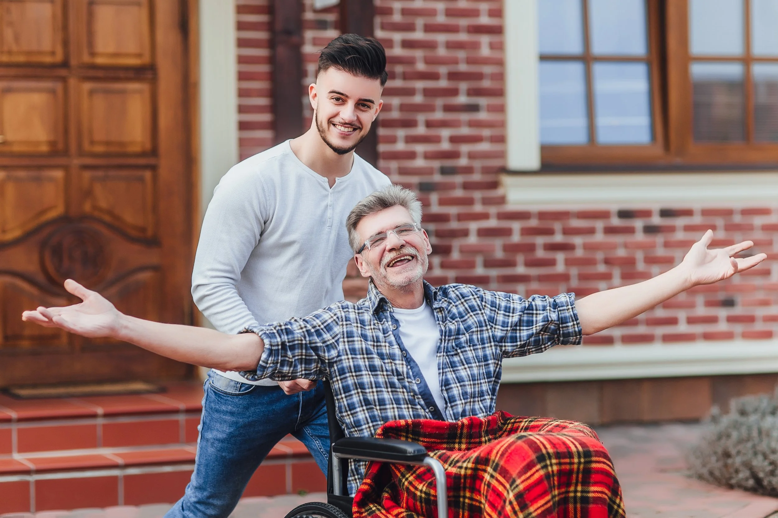 A young man pushing an older man in a wheelchair outside a brick house, both smiling and appearing joyful.